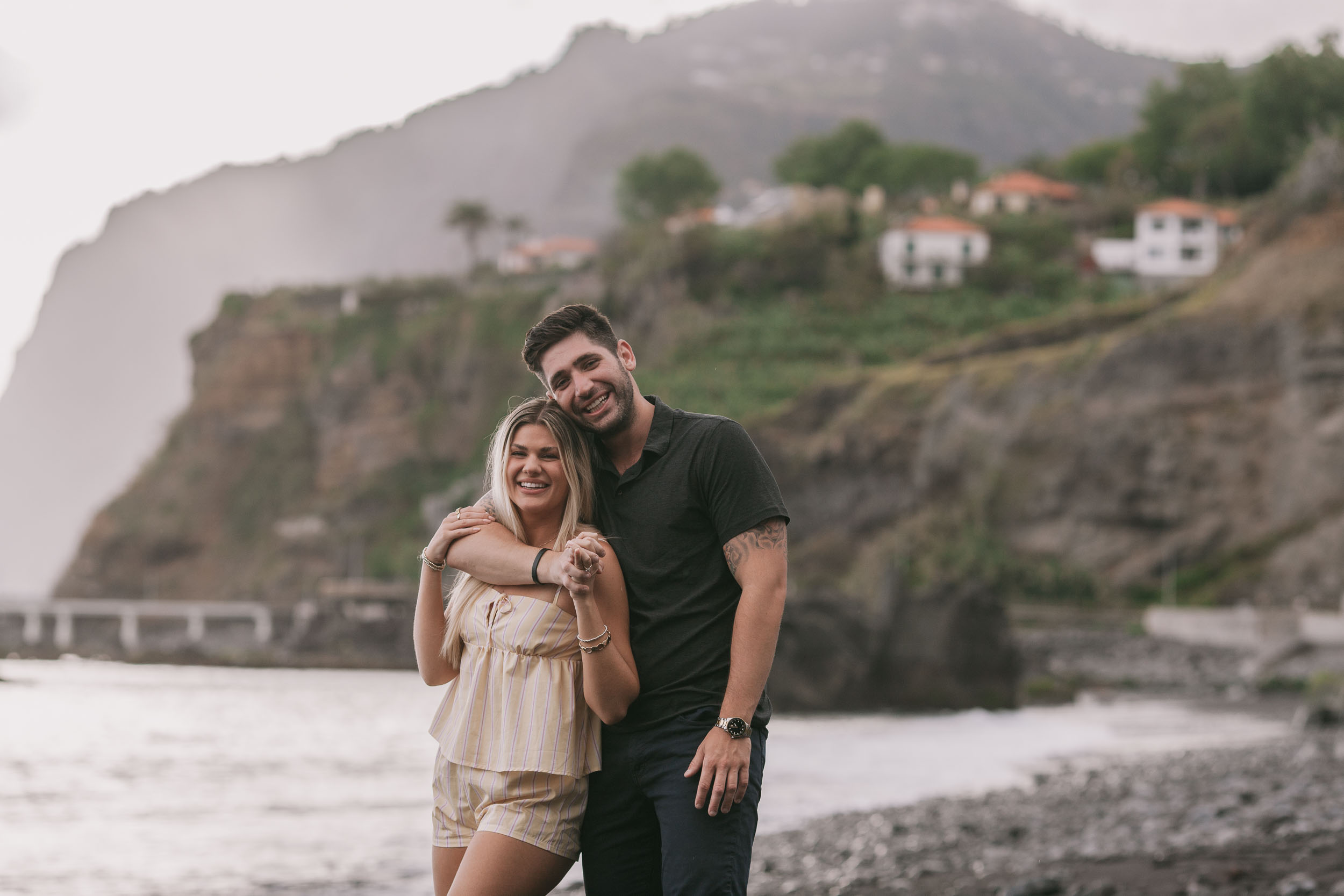 Mike standing behind Amelie with arms wrapped around her shoulders and his face beside hers smiling, the cliffside white houses and terraced gardens climbing the hill behind at Praia Formosa, Madeira