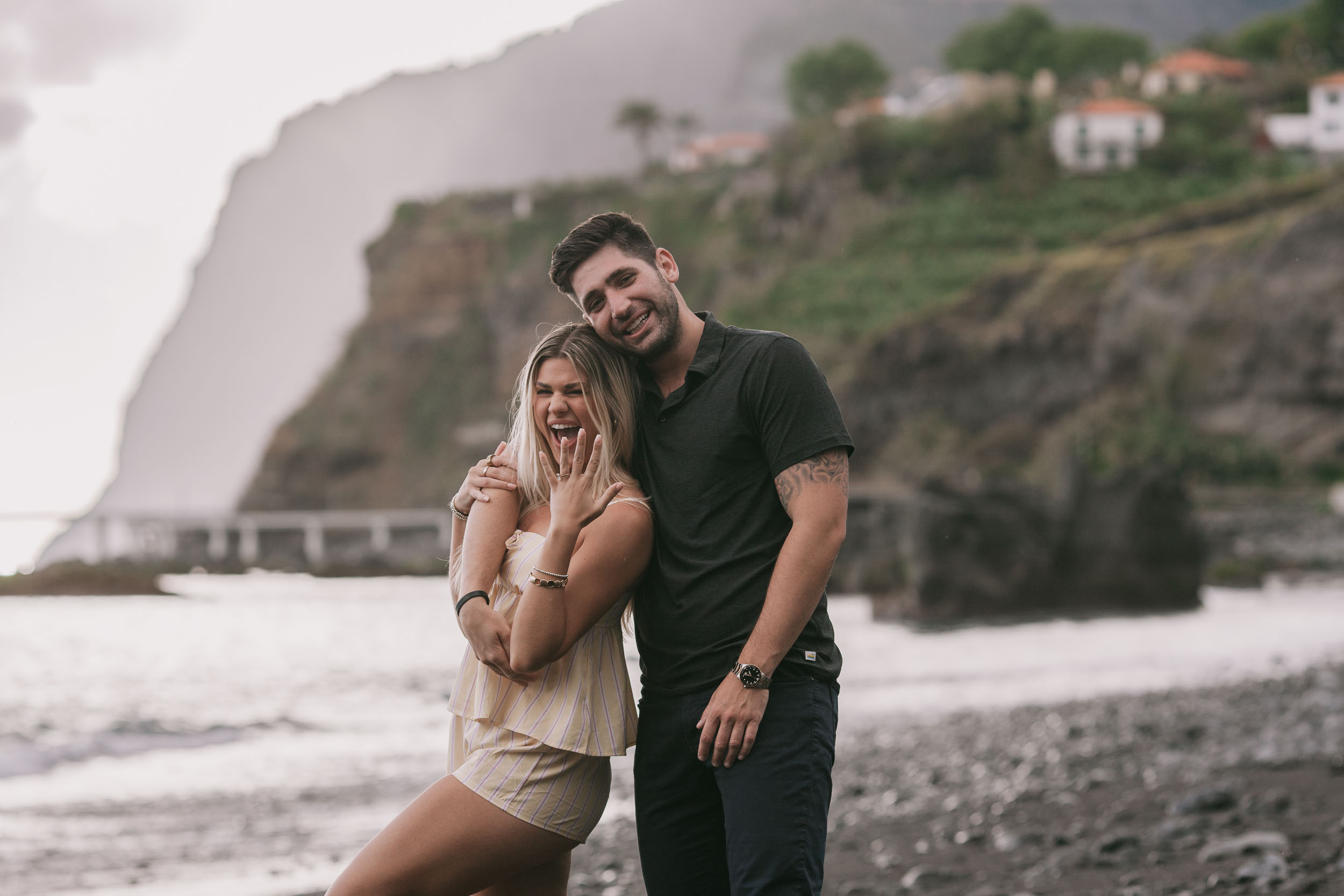 Amelie resting her cheek against Mike's chest with a wide laugh and closed eyes, his arm draped over her shoulders, terraced village houses and cliffs filling the background at Praia Formosa, Madeira