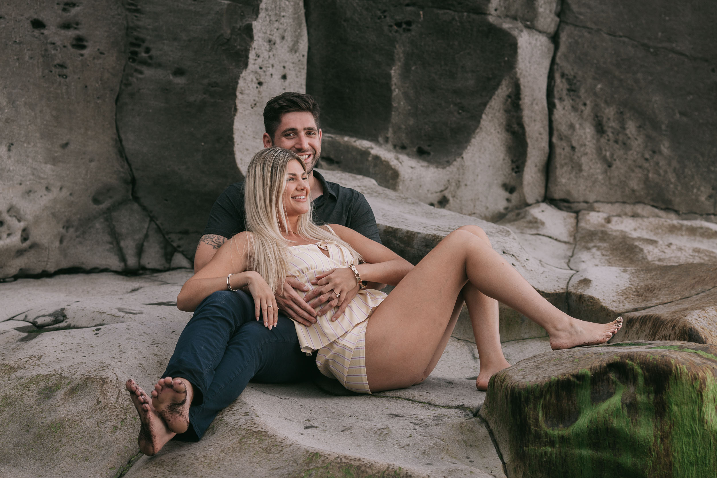 Mike sitting on a wide flat boulder with Amelie reclining back against his chest between his legs, both leaning back laughing, weathered grey volcanic rock wall filling the frame at Praia Formosa, Madeira