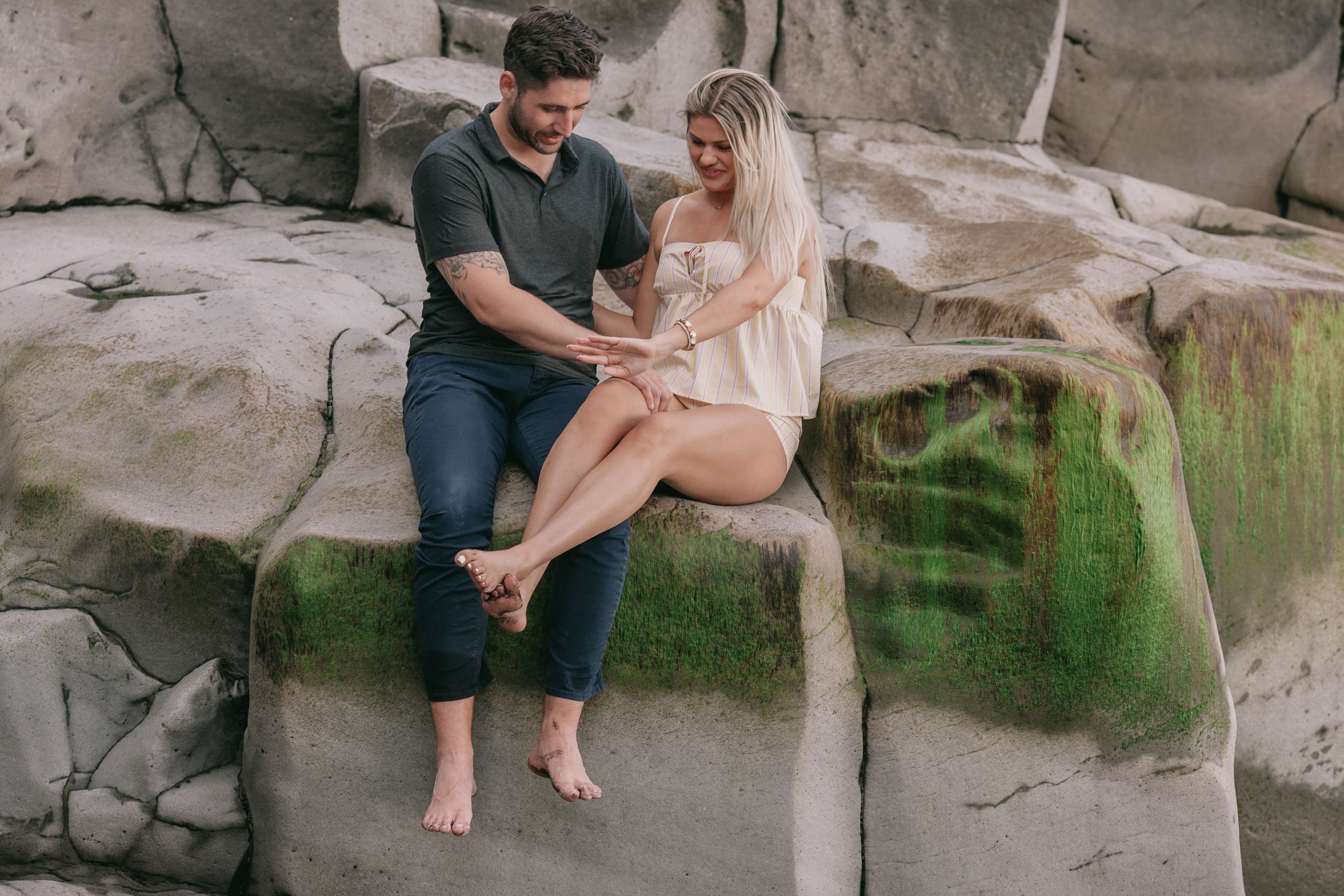 Mike and Amelie sitting beside each other on the flat rock shelf both looking down as he adjusts her bare foot resting on his knee, mossy green streaks running across the basalt at Praia Formosa, Madeira