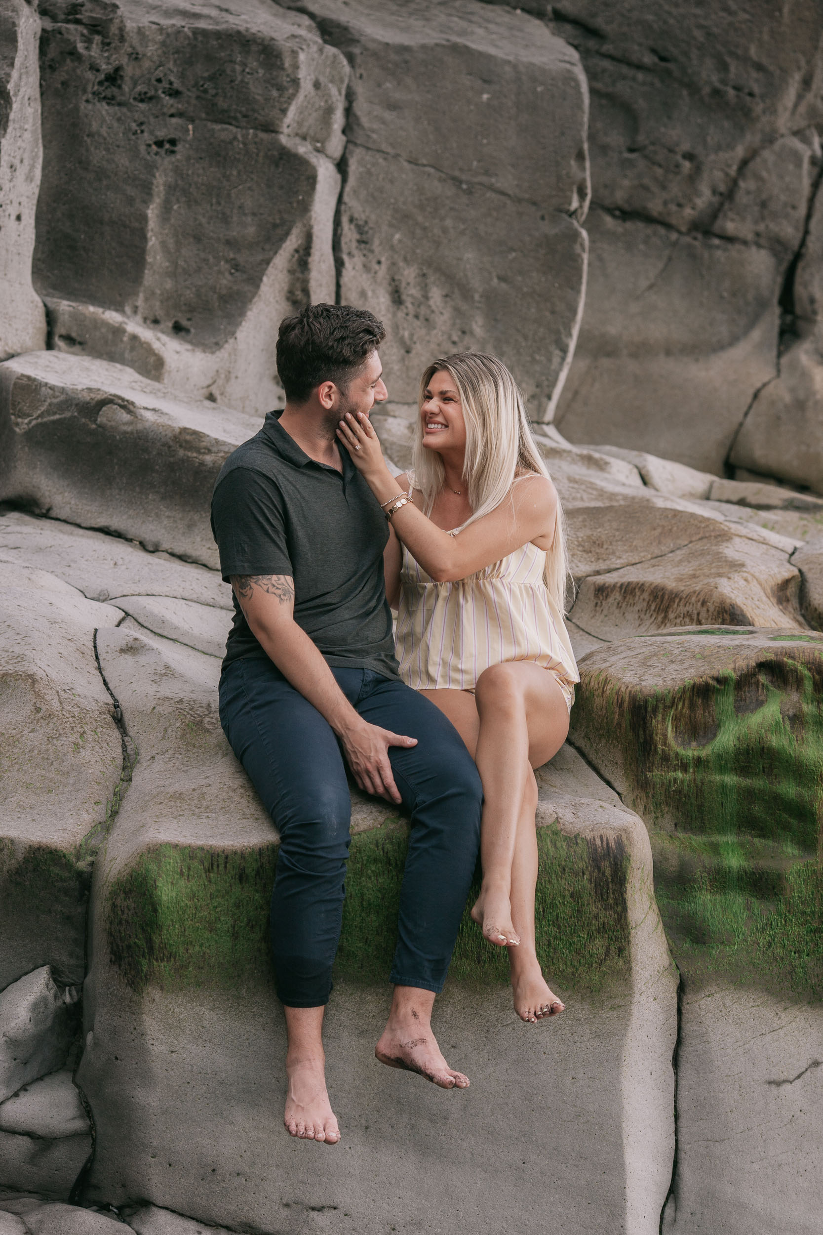 Amelie sitting on a wide flat boulder with her bare legs across Mike's lap, one hand reaching up to rest on his cheek as she smiles up at him, mossy basalt around them at Praia Formosa, Madeira