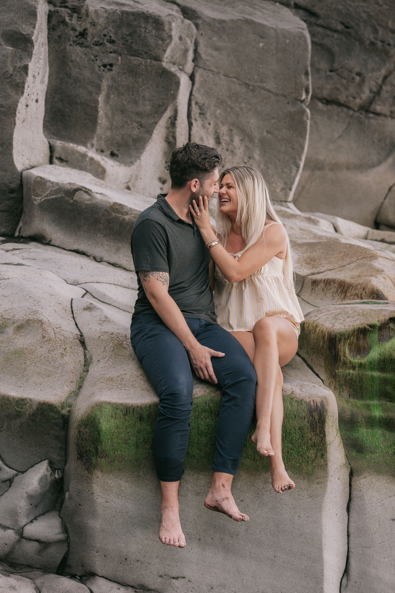 Mike and Amelie sitting side by side on the rock shelf laughing nose to nose, her ringed hand resting on his jaw, his tattooed forearm visible, moss-green streaks across the stone at Praia Formosa, Madeira
