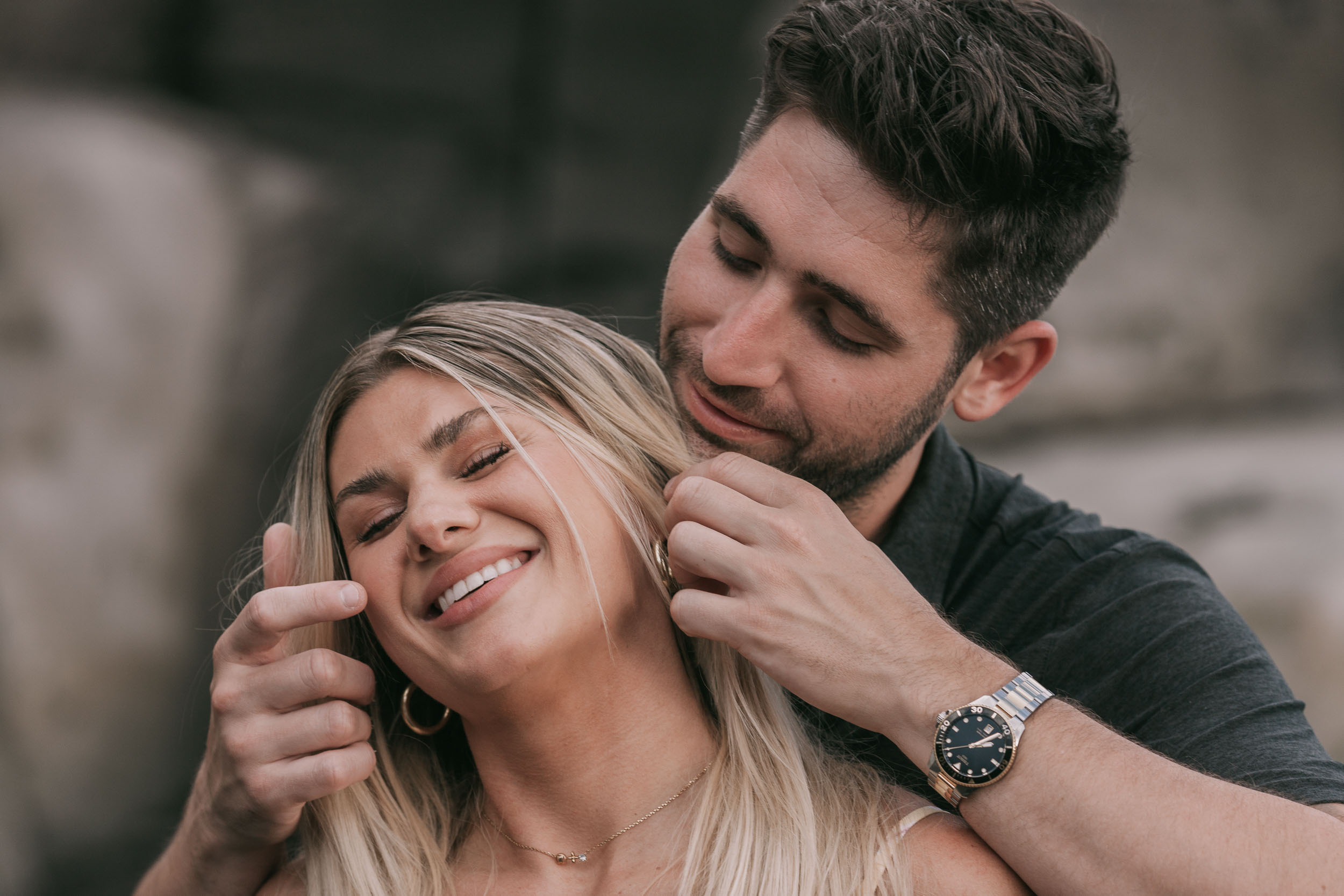 Close portrait of Mike gently tucking Amelie's hair behind her ear from behind while she laughs and lifts a hand in mock protest, his silver-and-gold watch on his wrist at Praia Formosa, Madeira