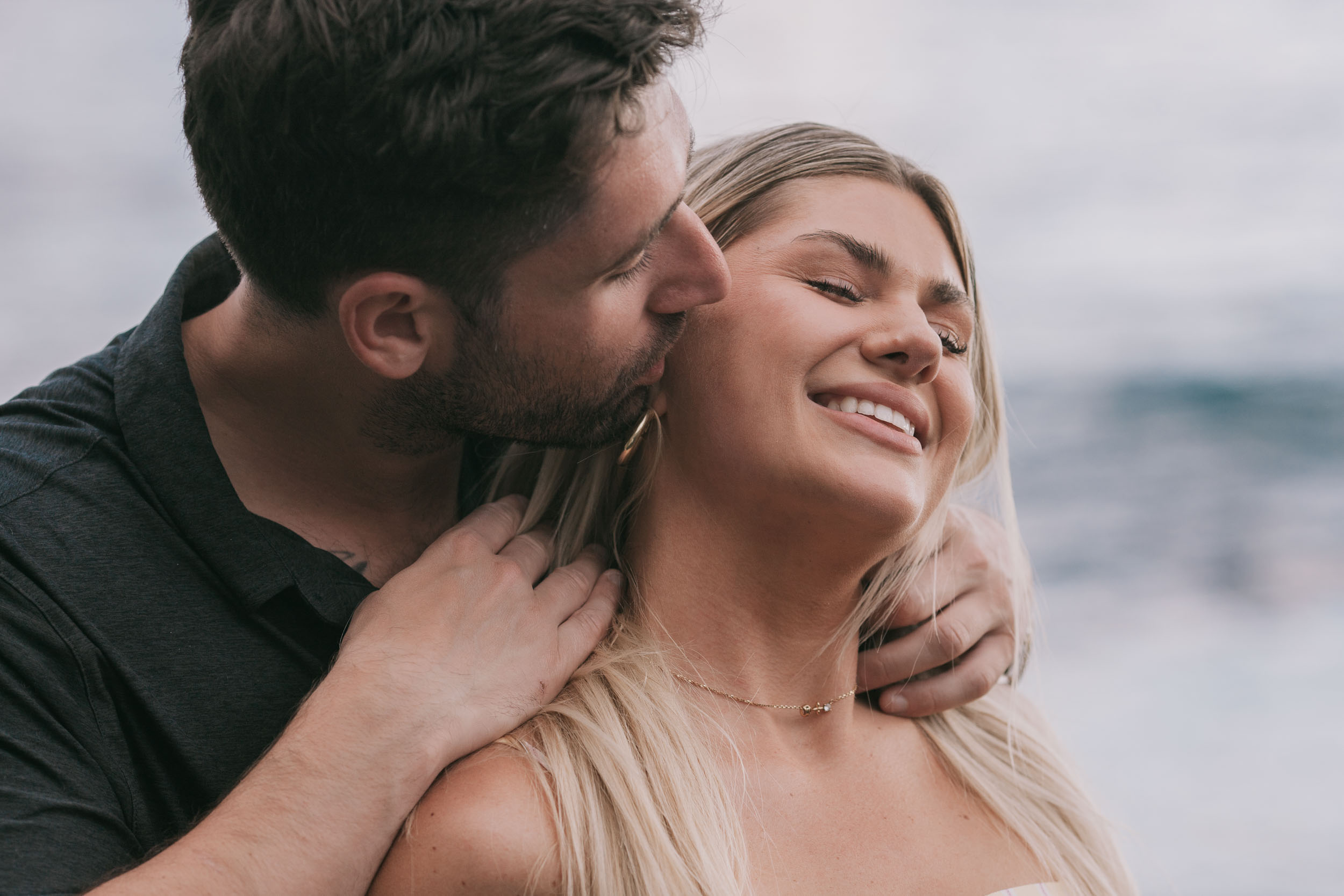 Tight close portrait of Mike kissing Amelie's cheek from the side as her face tips up smiling with eyes closed, soft blue water blurred behind at Praia Formosa, Madeira