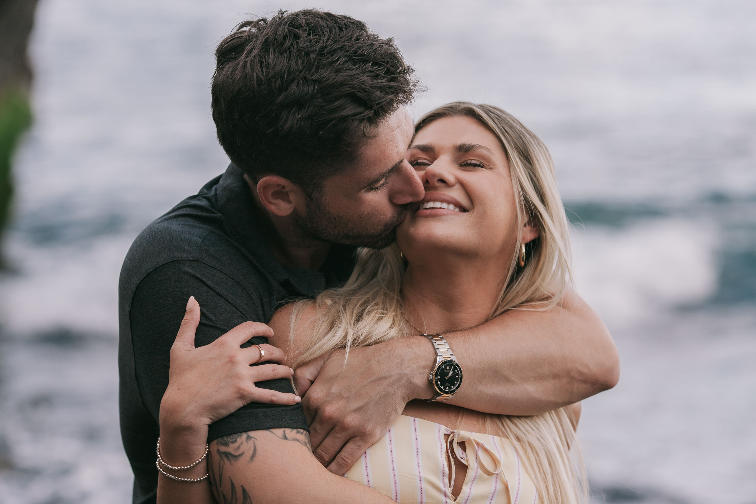 Mike kissing Amelie's cheek with his arms folded tightly across her chest from behind, her wide grin and his bracelet and tattoo sleeve visible, soft white Atlantic behind at Praia Formosa, Madeira