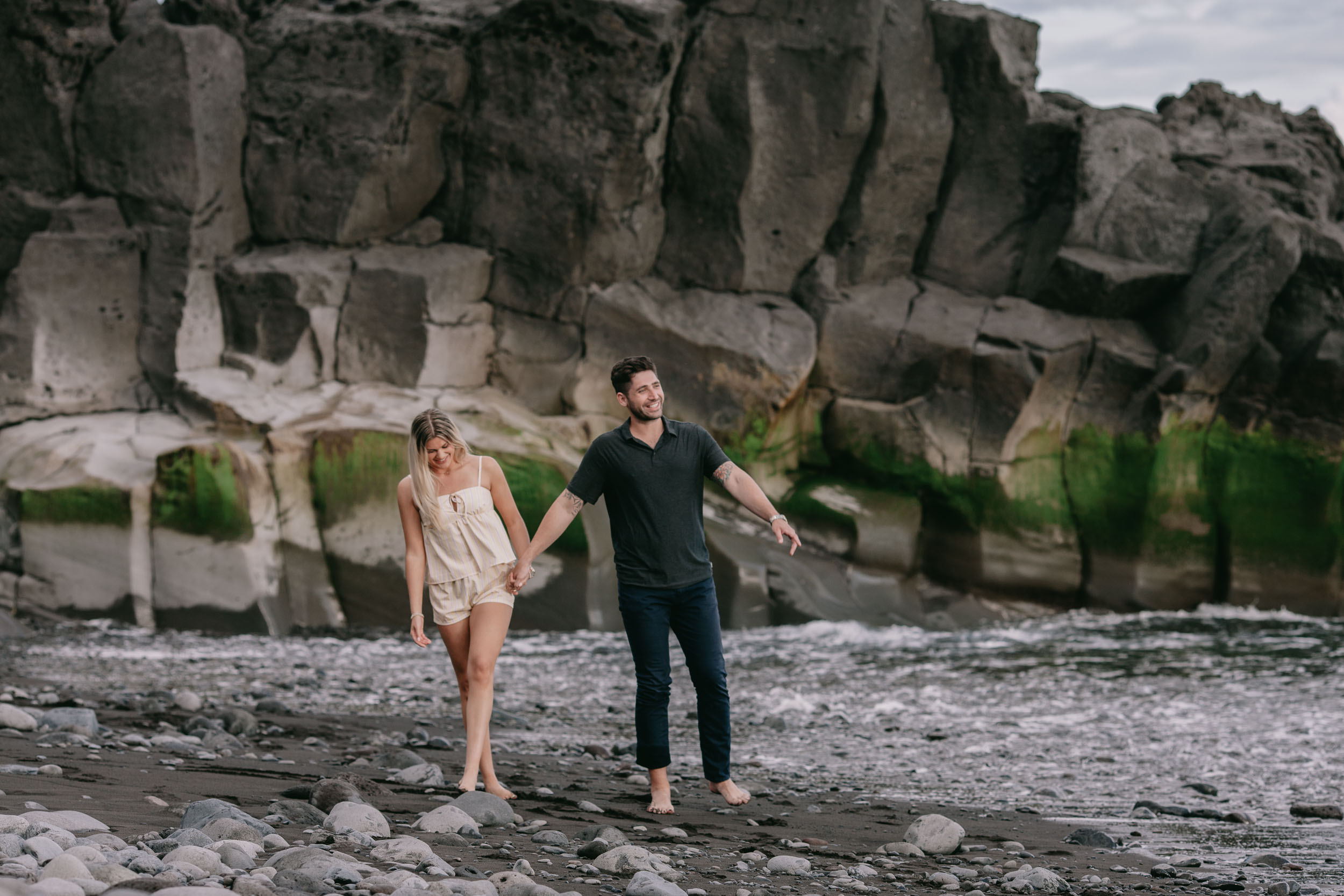 Mike and Amelie walking out together from below a tall mossy basalt rock wall onto the pebble shore, white wave breaking on the right at Praia Formosa, Madeira