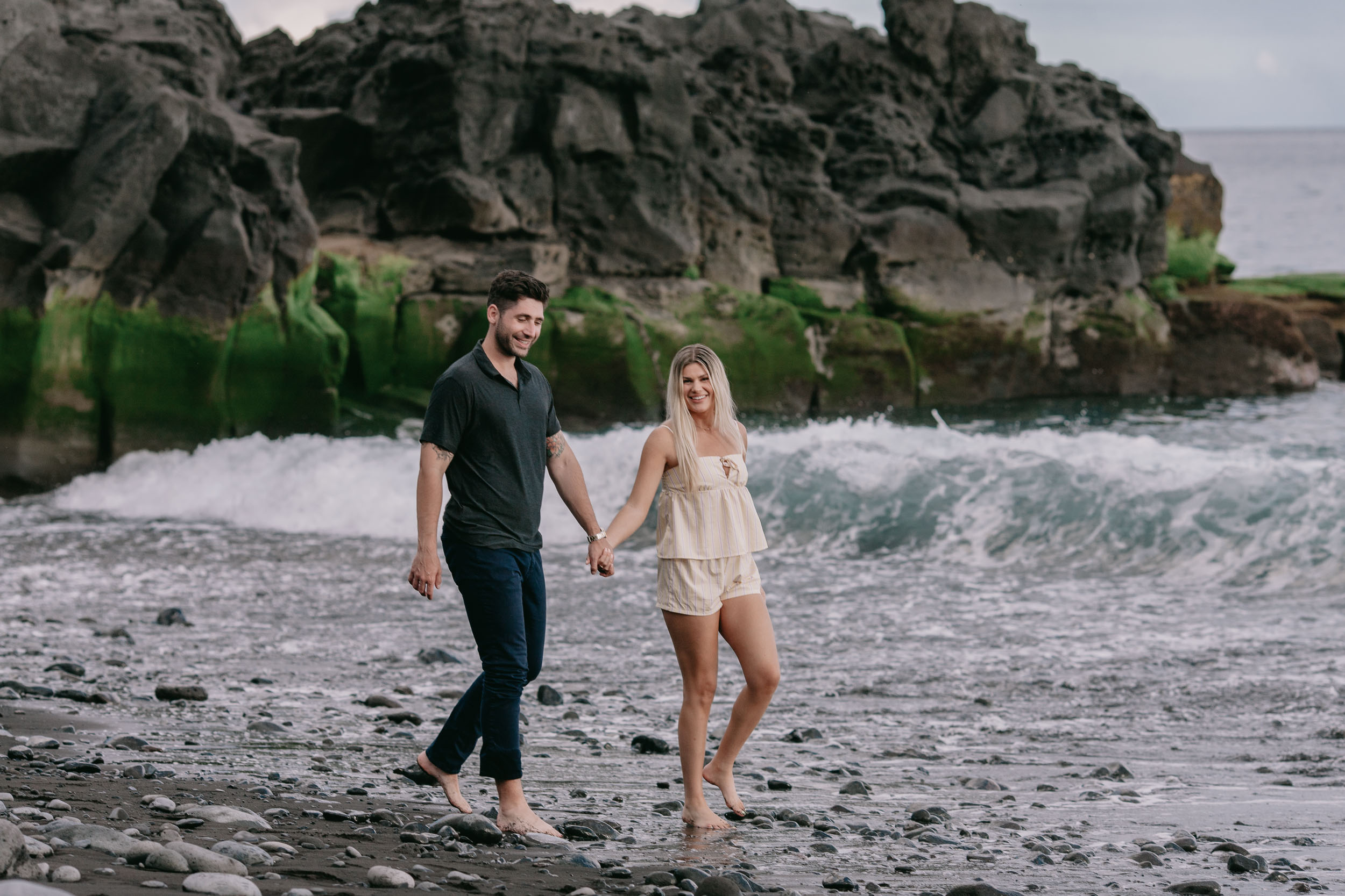 Mike and Amelie holding hands as they walk past a curved volcanic rock arch streaked with bright green algae, a curling teal wave breaking just beside them at Praia Formosa, Madeira