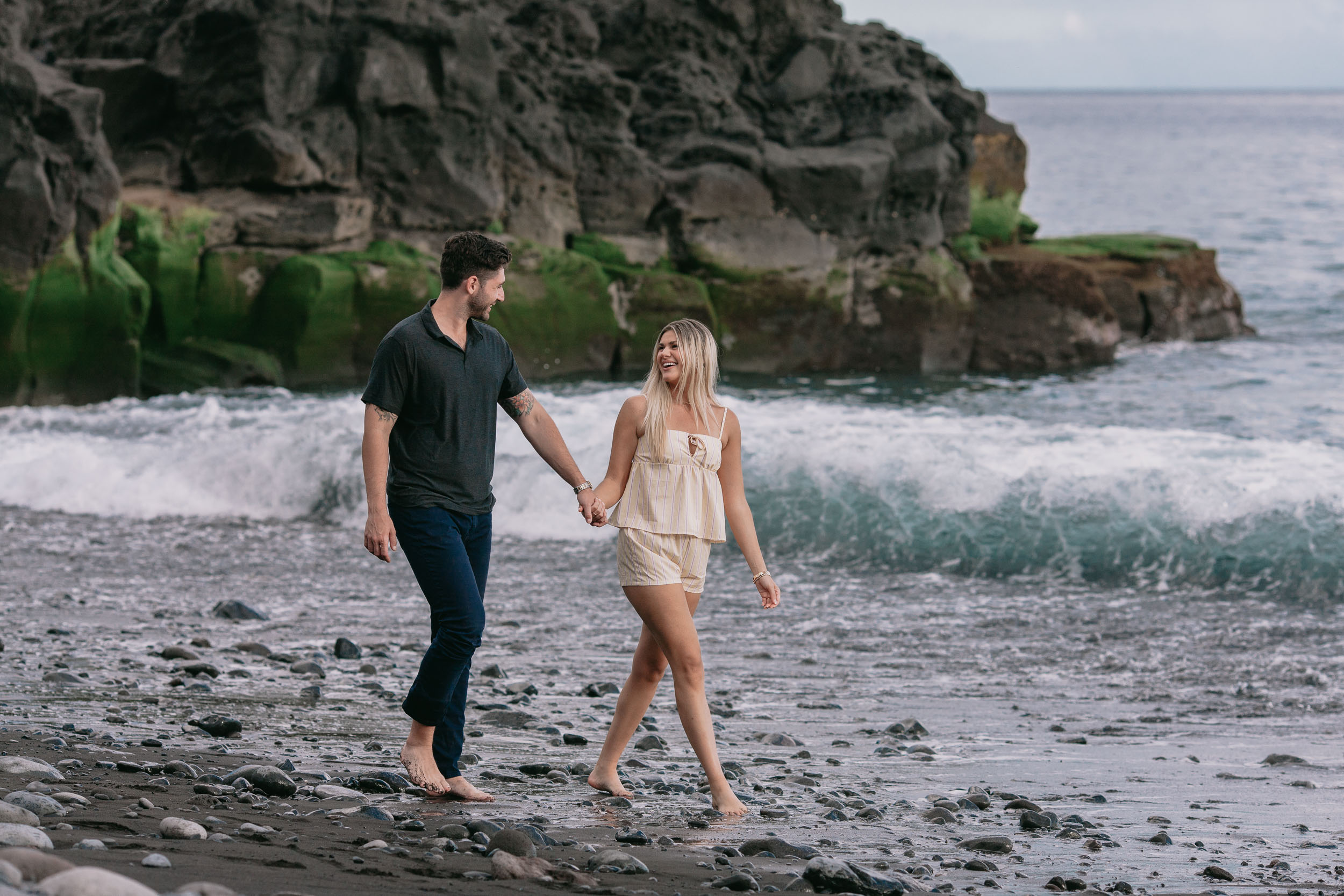 Mike and Amelie holding hands walking toward the camera below a tall mossy basalt outcrop, a curling wave folding over behind them at Praia Formosa, Madeira