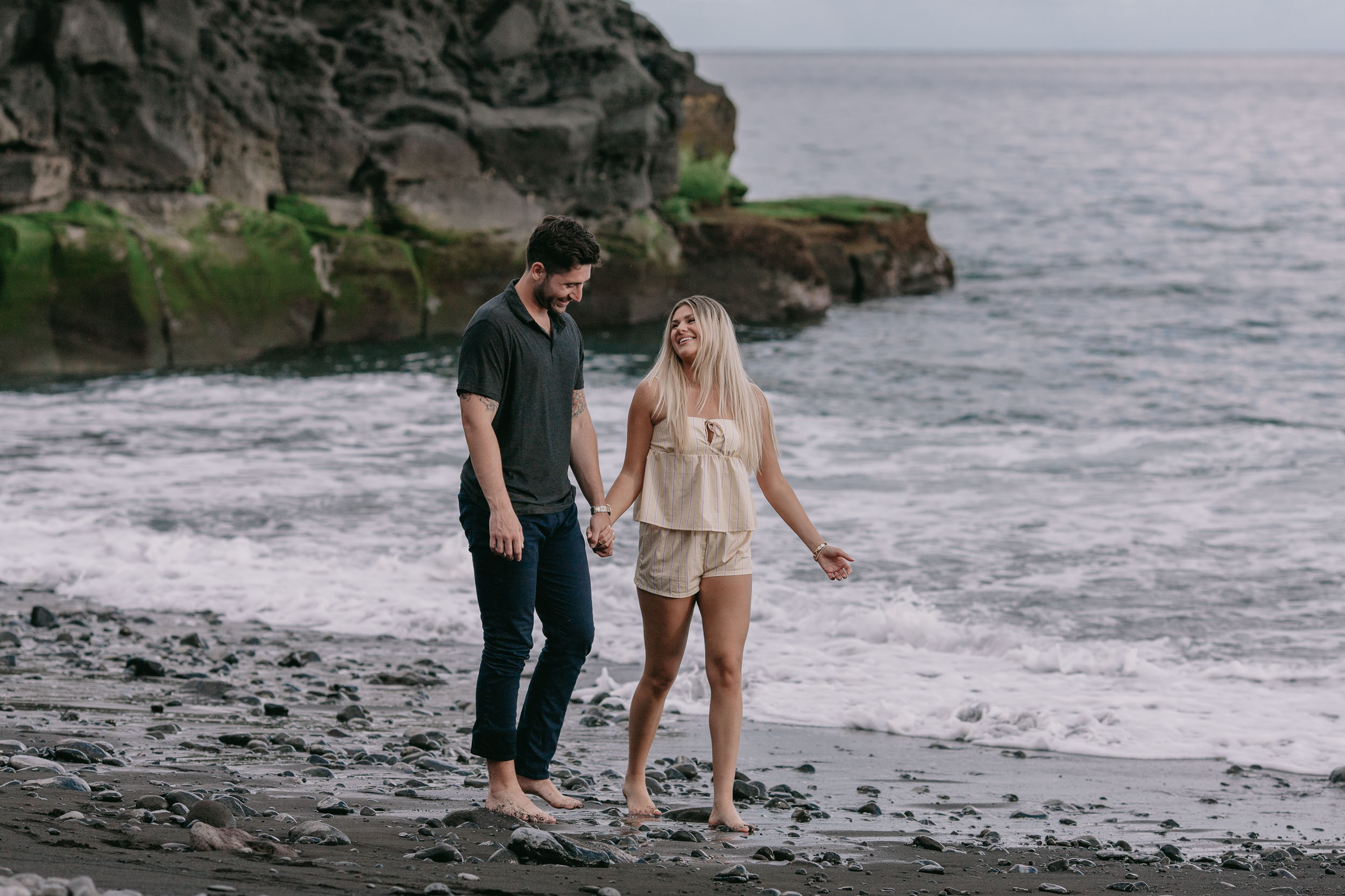 Mike and Amelie walking toward the camera barefoot at the shoreline, the small mossy headland on the left and bright ocean behind them at Praia Formosa, Madeira