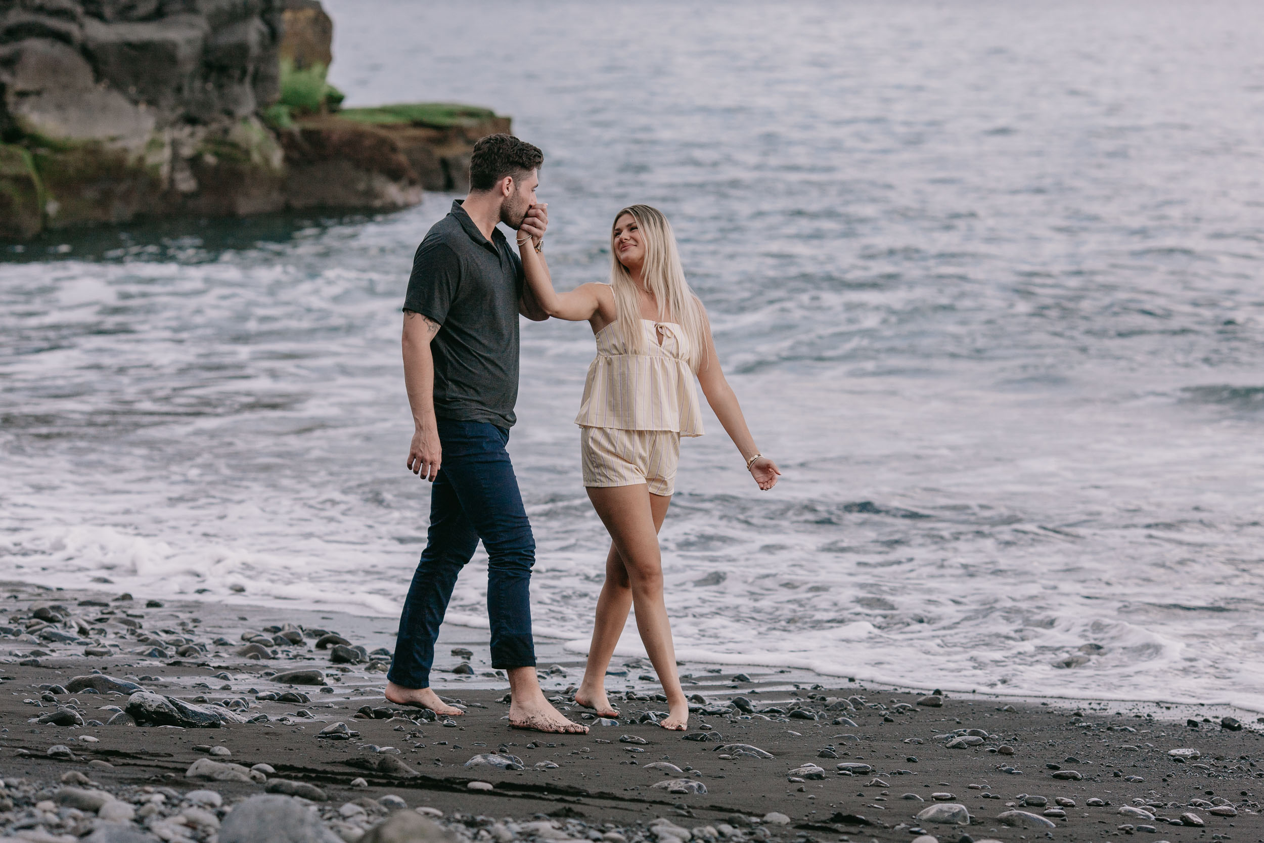 Mike bringing Amelie's hand up to kiss her knuckles as they walk along the dark sand, the small mossy headland and soft Atlantic water behind at Praia Formosa, Madeira
