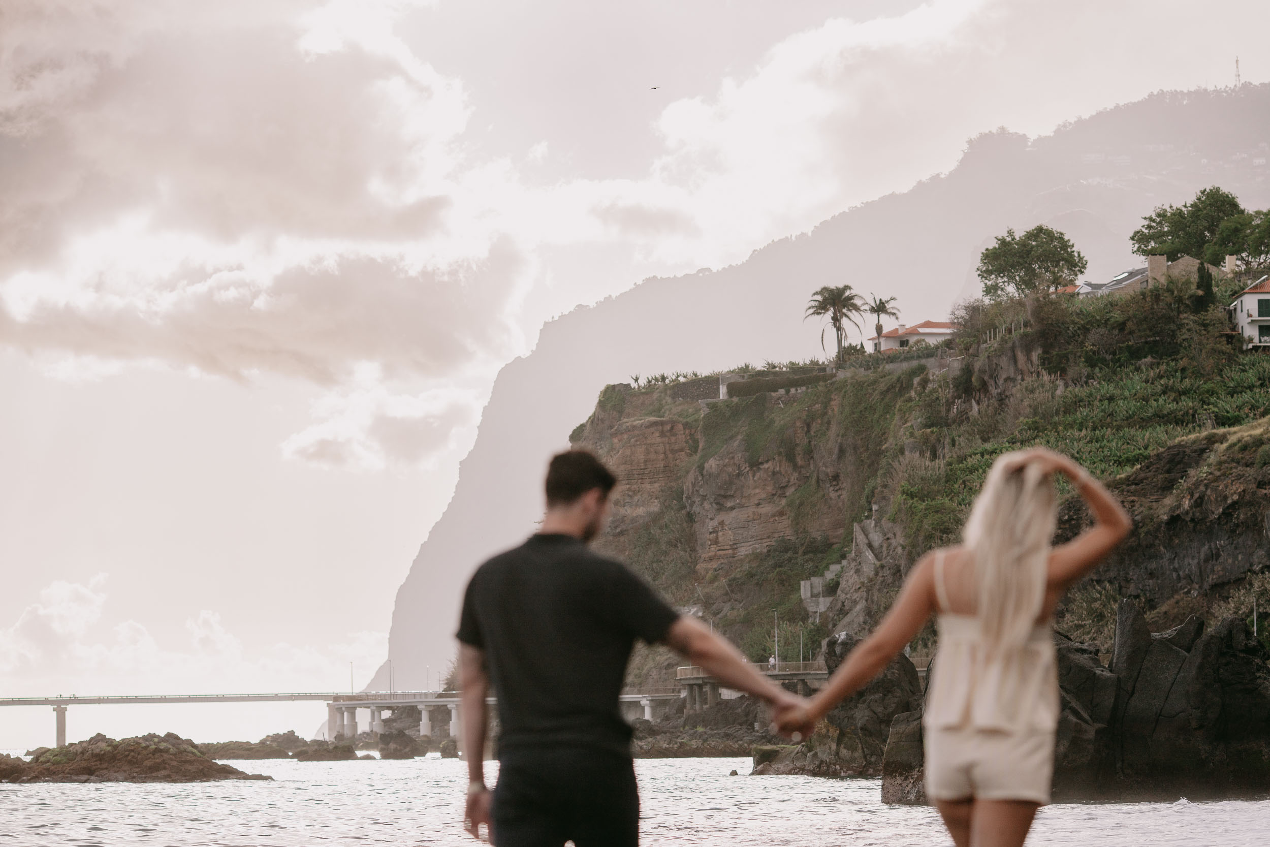 Mike and Amelie seen from behind standing in the shallows holding hands, both gazing out at the green Cabo Girão cliff and the long coast road viaduct in the distance at Praia Formosa, Madeira