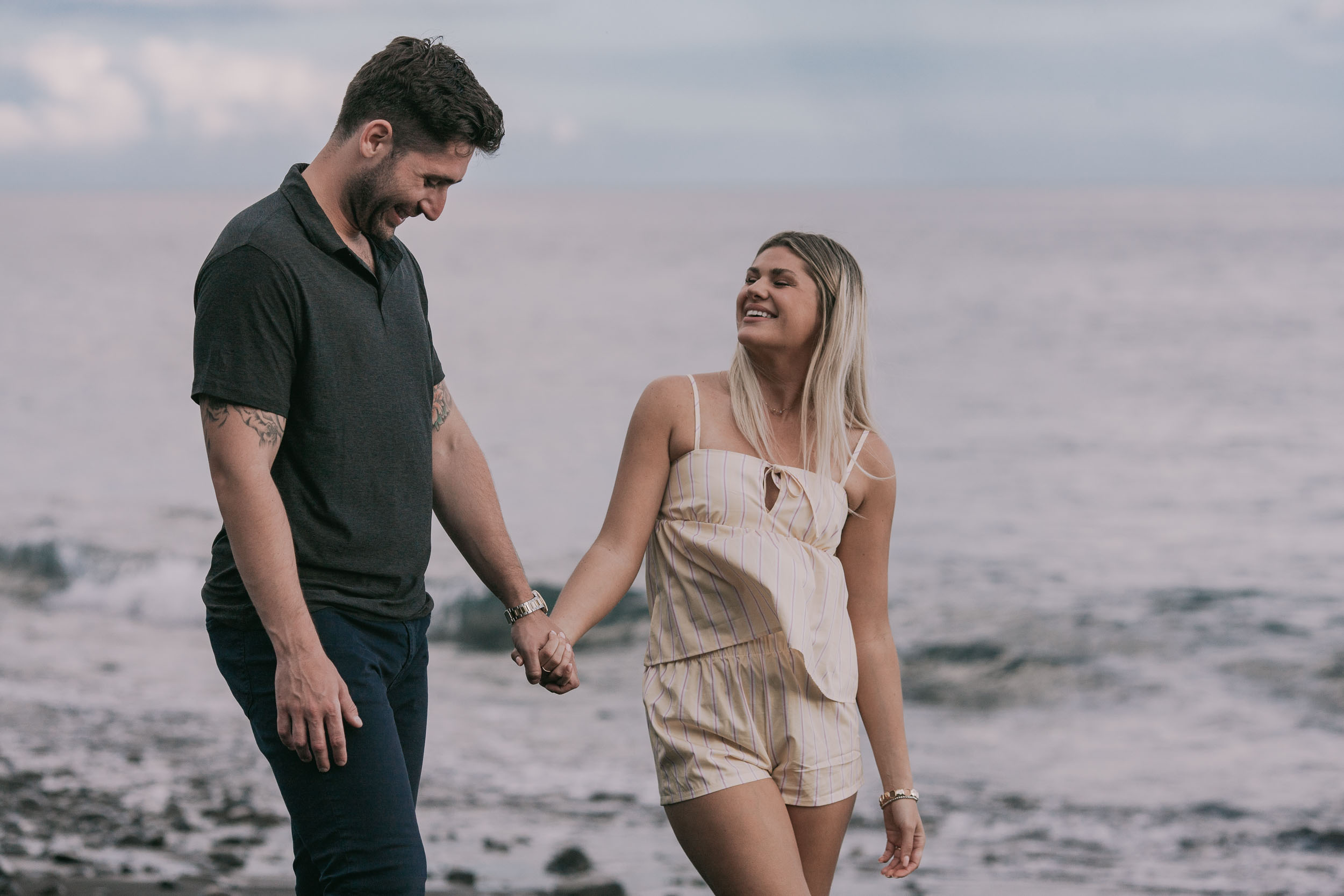 Amelie looking up at Mike laughing as they walk close together holding hands, her ring just visible, soft grey Atlantic horizon behind them at Praia Formosa, Madeira