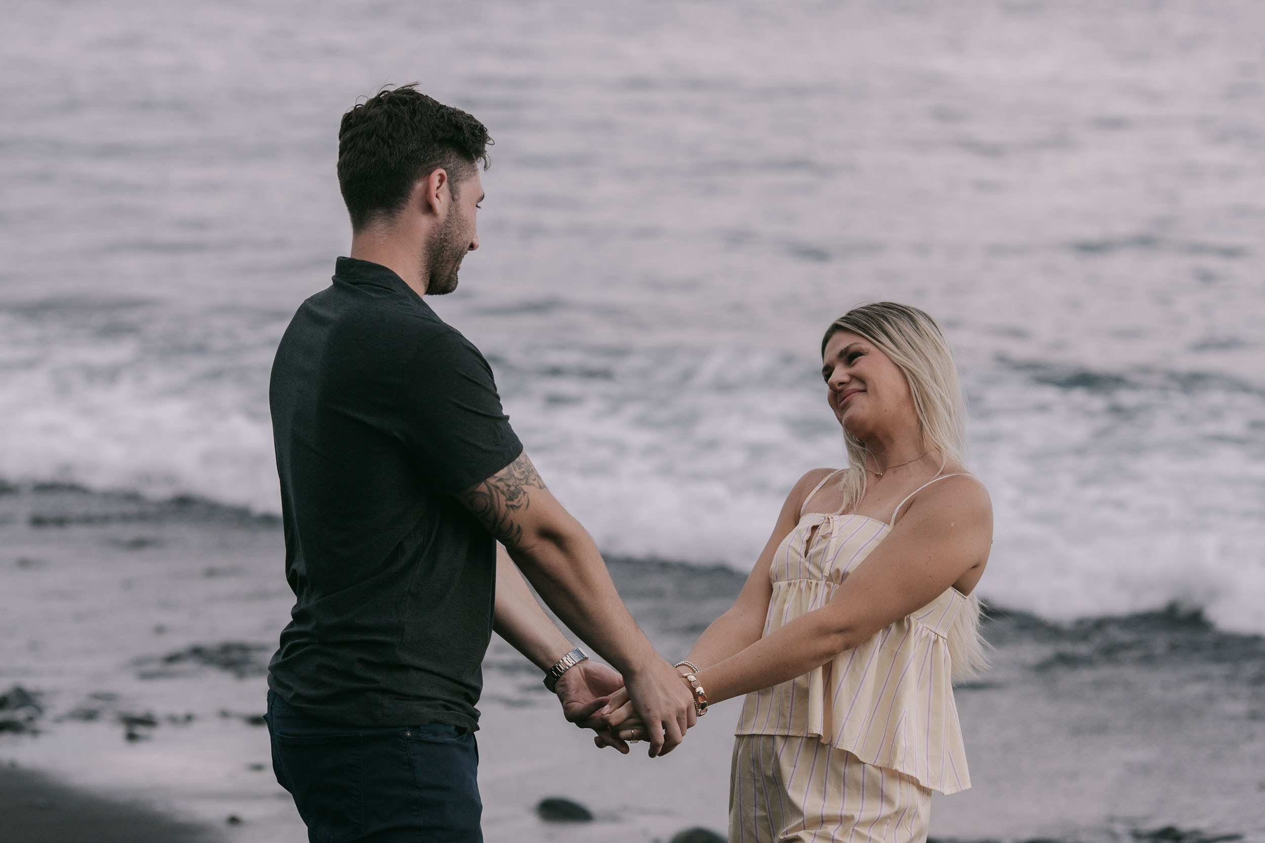 Mike and Amelie standing facing each other on the dark sand with both hands clasped between them, her face crumpled in a happy laugh, foamy surf behind at Praia Formosa, Madeira