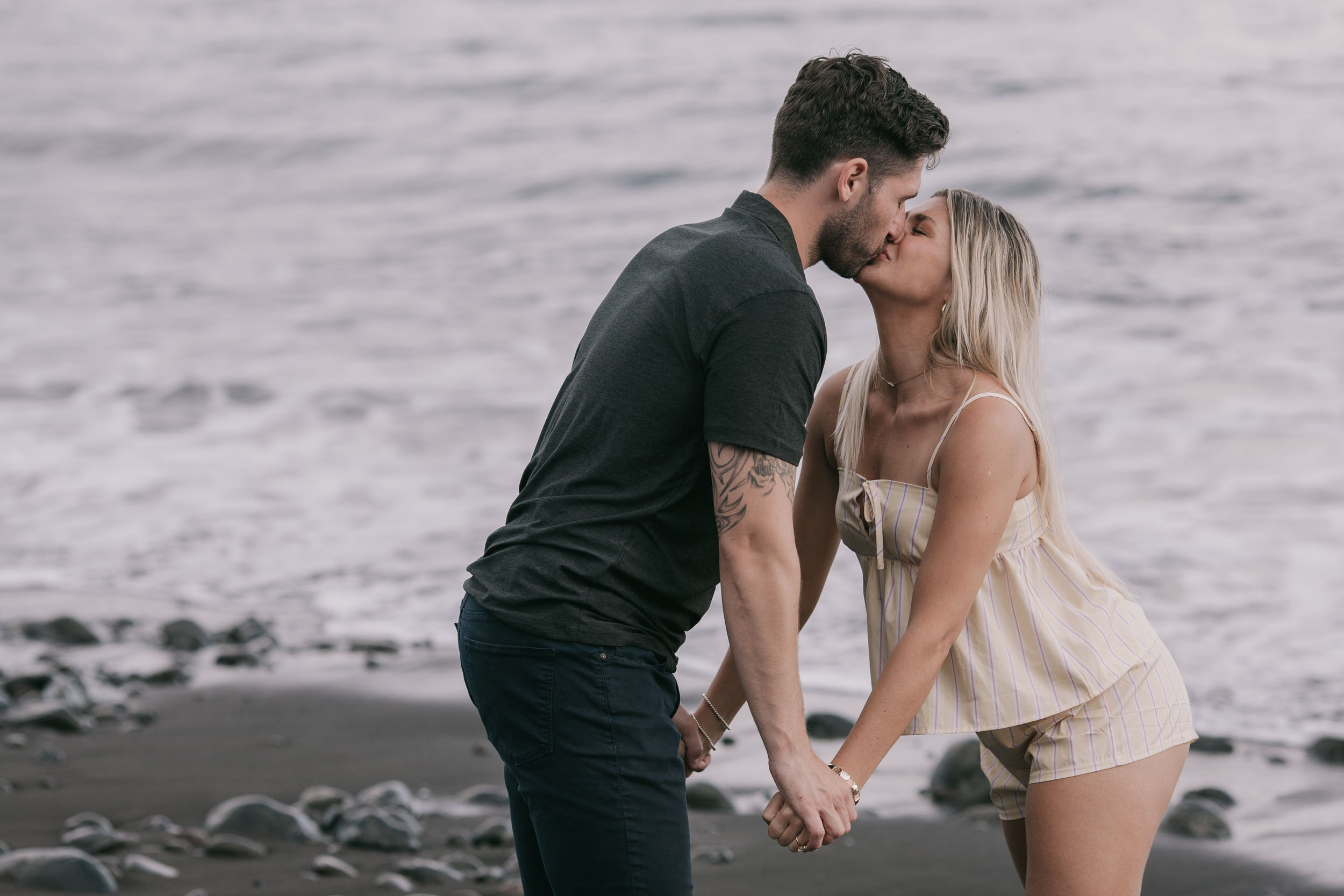 Mike leaning in to kiss Amelie as they stand holding hands at the shoreline, both bowed slightly toward each other, foam rushing past their feet at Praia Formosa, Madeira