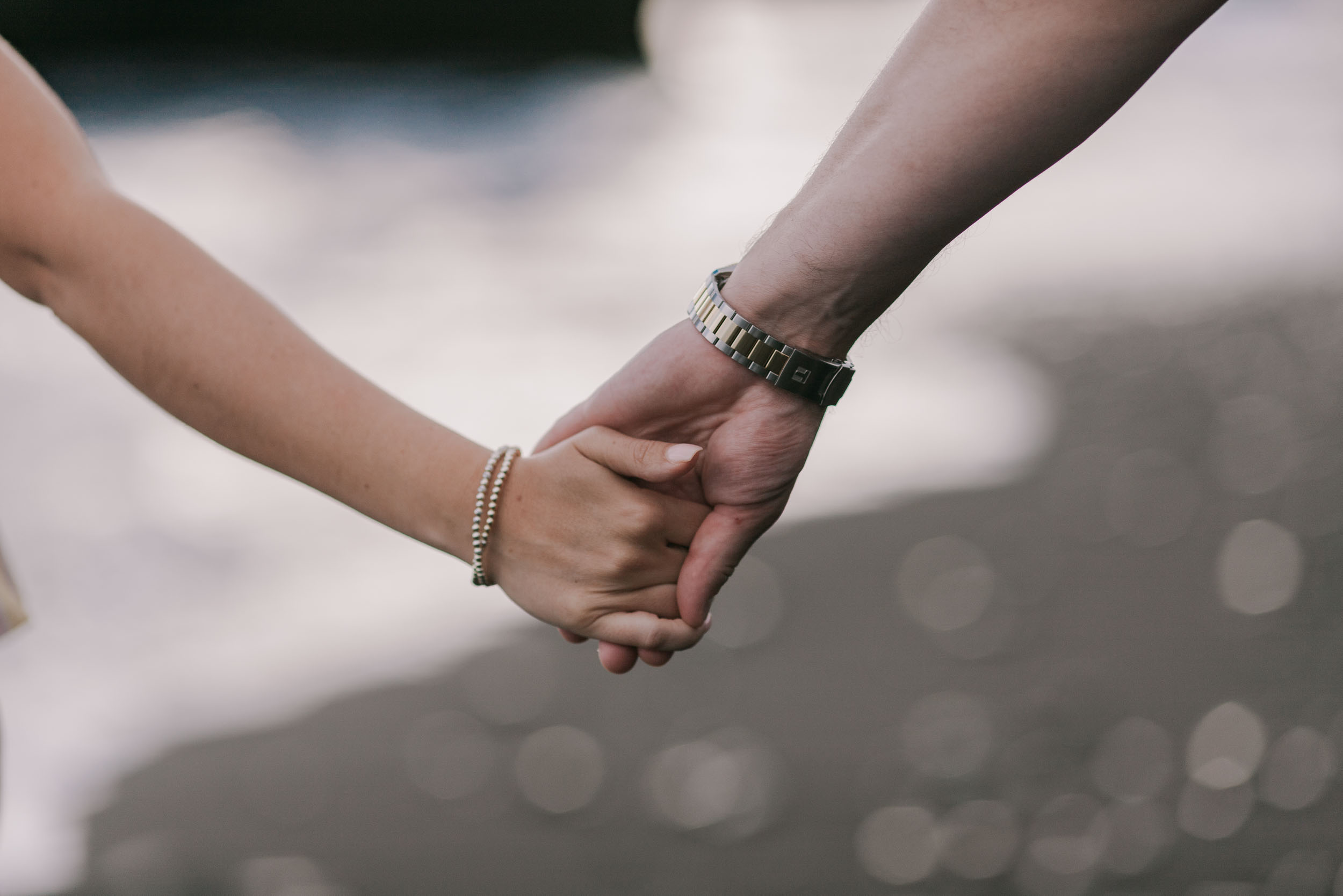 Tight close-up of Mike's and Amelie's hands fully clasped at chest level, his metal watch wrapping his wrist and her thin gold bracelets stacked on hers, soft bokeh of surf behind at Praia Formosa, Madeira