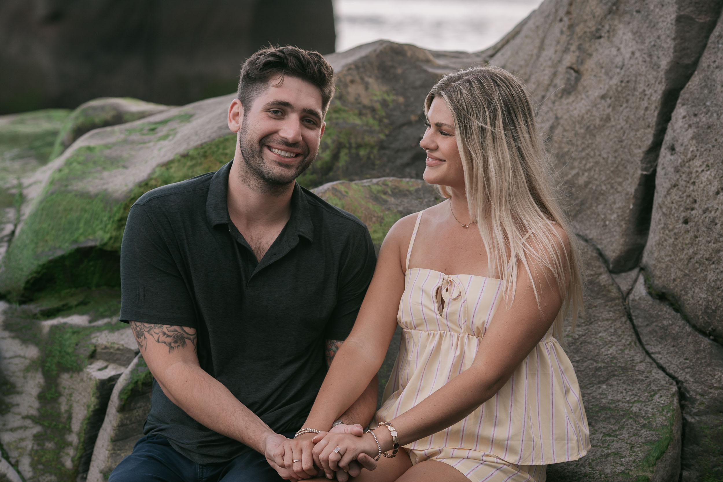Mike and Amelie sitting tucked between two large mossy boulders with hands joined in their laps, both smiling straight at the camera, Praia Formosa, Madeira