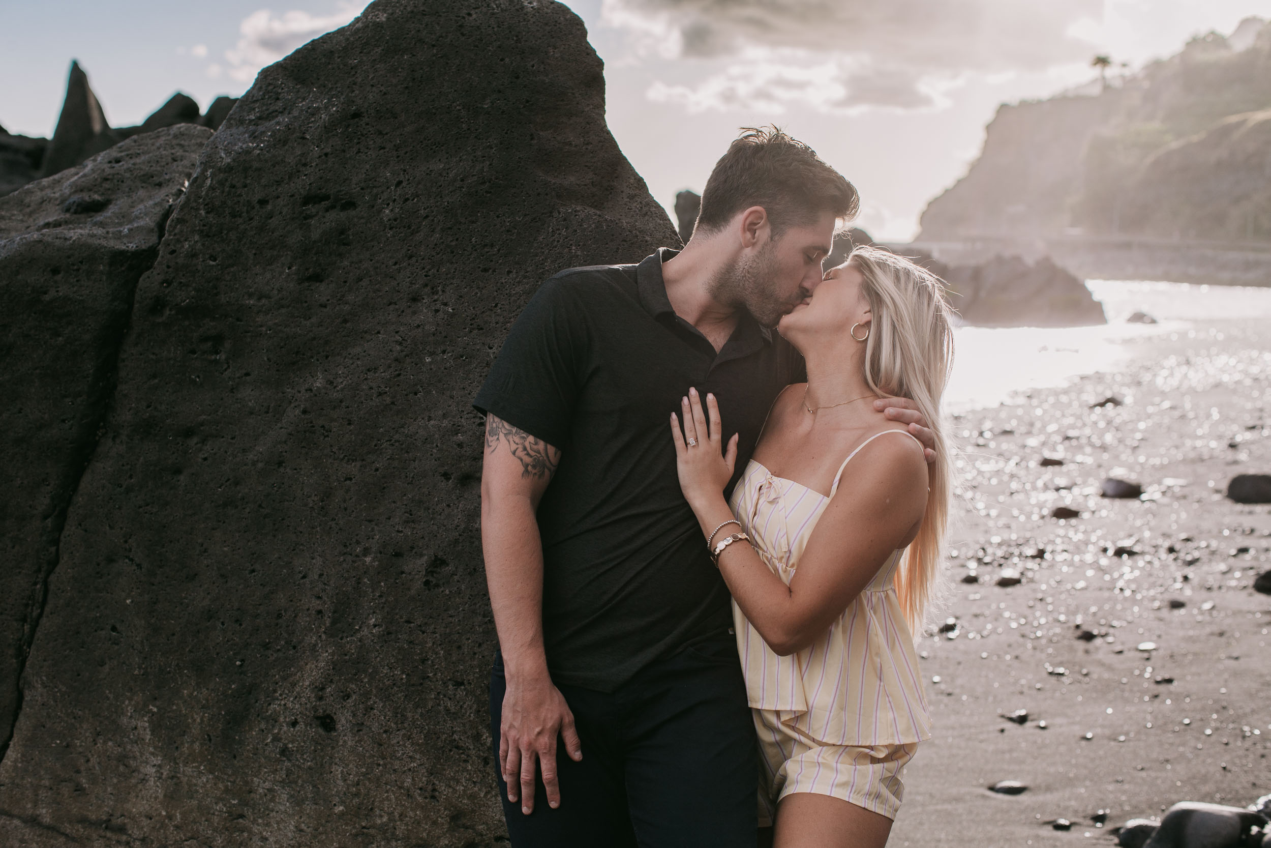 Mike and Amelie kissing standing pressed against a tall black volcanic boulder, her hand resting on his chest, warm sun breaking under the clouds at the horizon at Praia Formosa, Madeira
