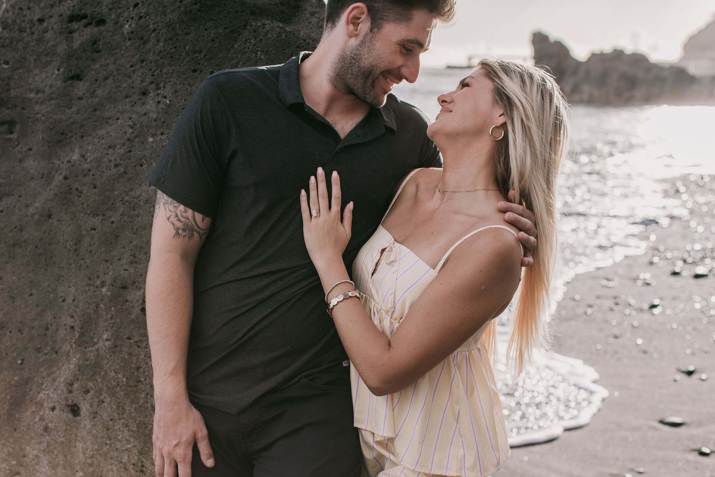 Amelie smiling up at Mike with her hand on his chest as he grins beside the tall basalt rock, soft afternoon shimmer on the wet sand behind at Praia Formosa, Madeira