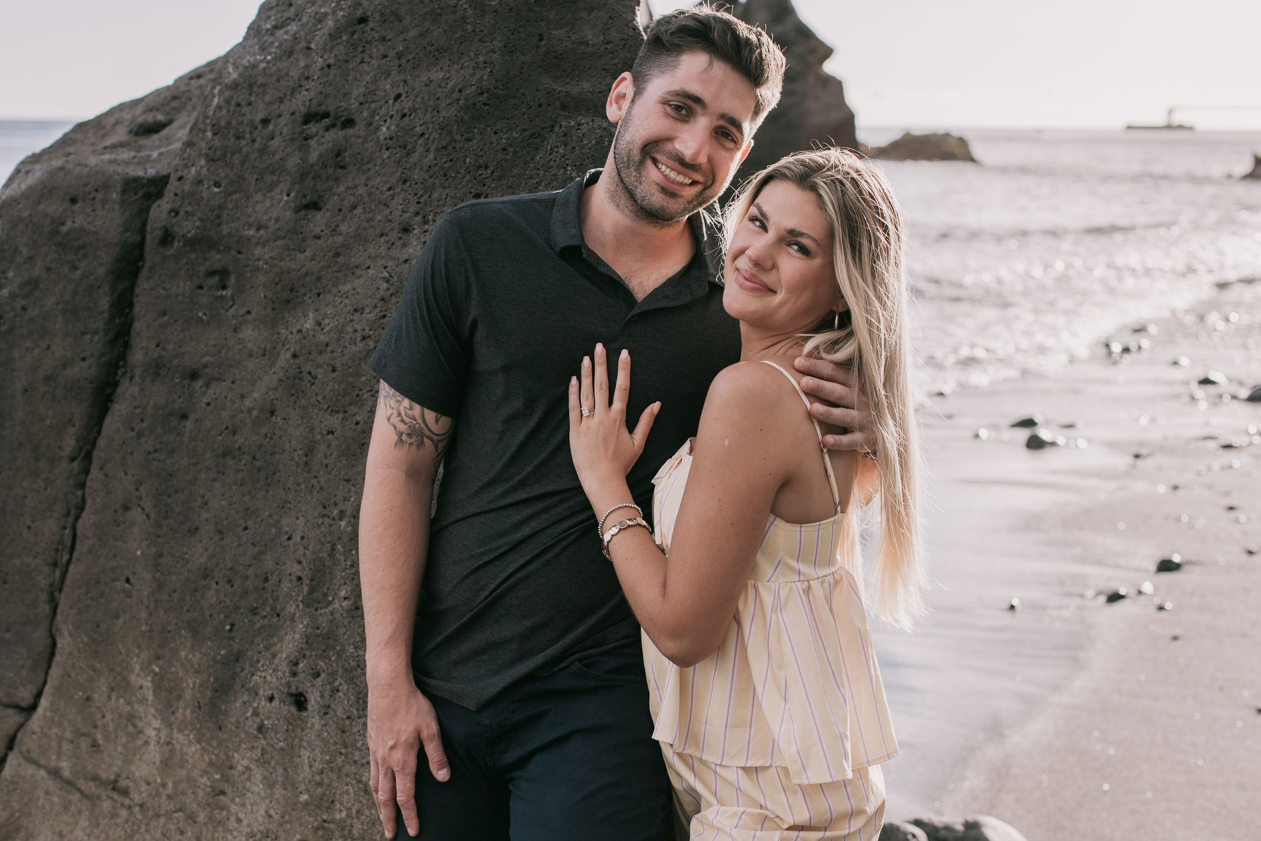 Mike and Amelie both turned to smile at the camera while leaning against a large black volcanic boulder, her ringed hand on his chest, glittering wet sand behind at Praia Formosa, Madeira