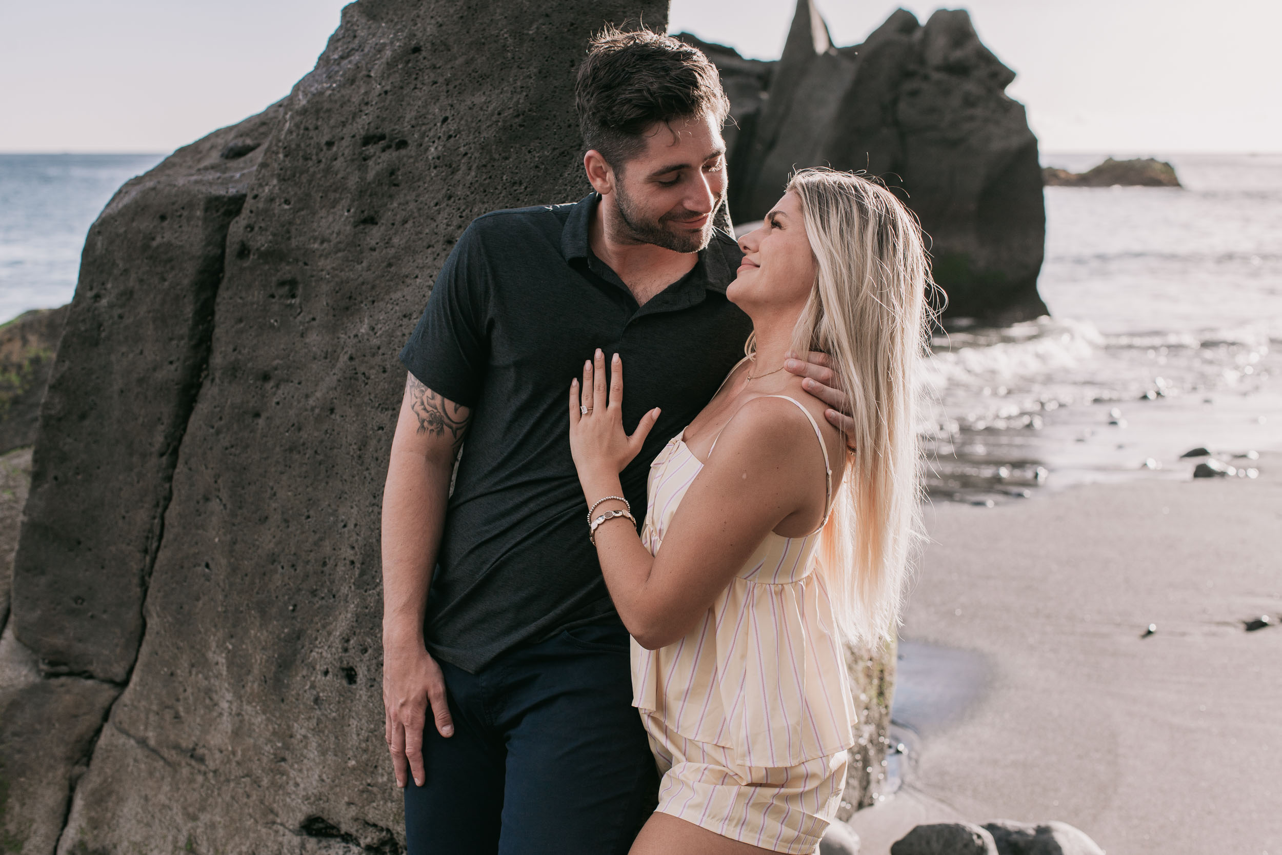 Amelie's ringed hand flat on Mike's chest as she gazes up at him laughing softly, both leaning against the dark volcanic rock face at Praia Formosa, Madeira
