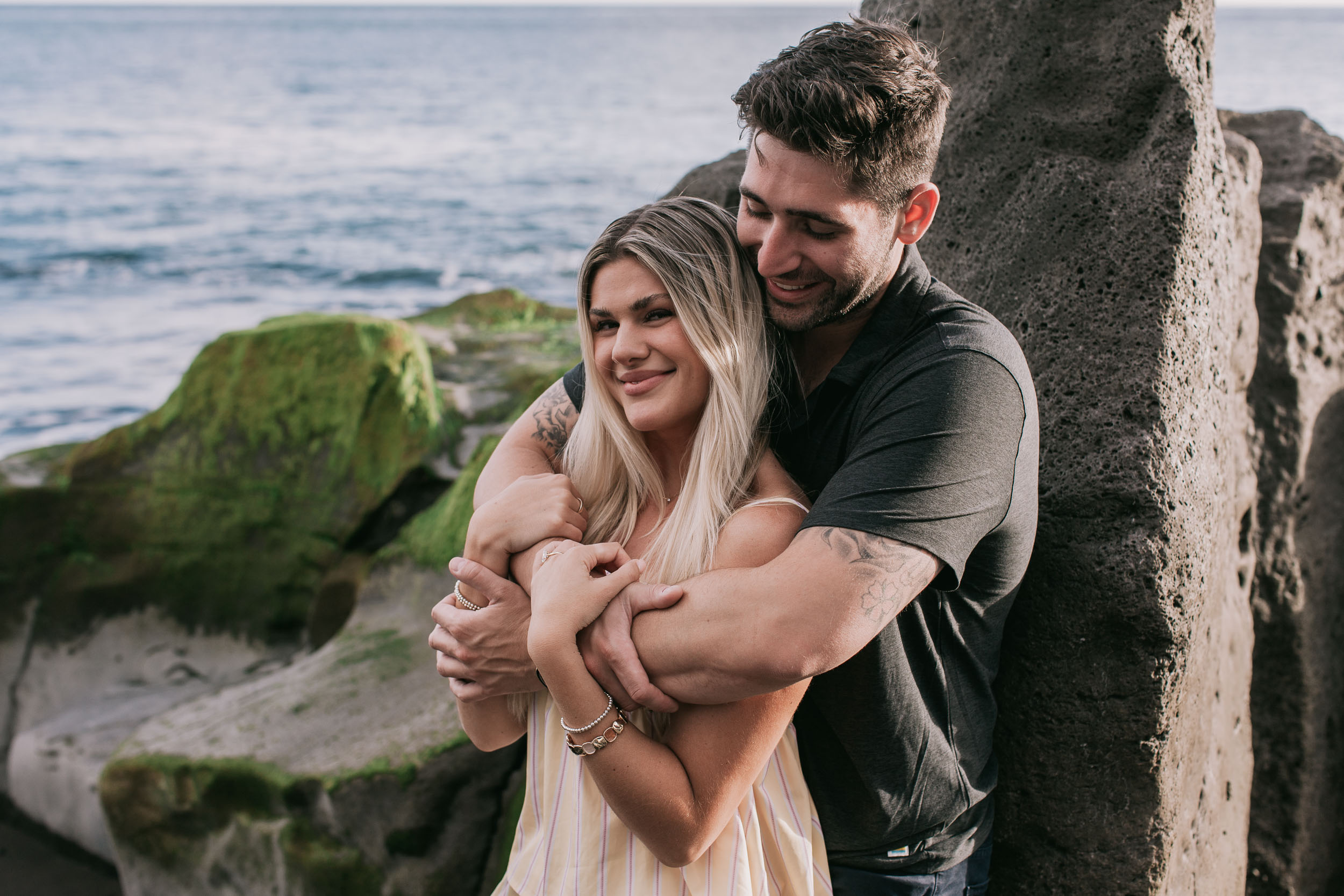 Mike hugging Amelie from behind in front of a moss-streaked black cliff face, both smiling with half-closed eyes, blue Atlantic visible on the left at Praia Formosa, Madeira