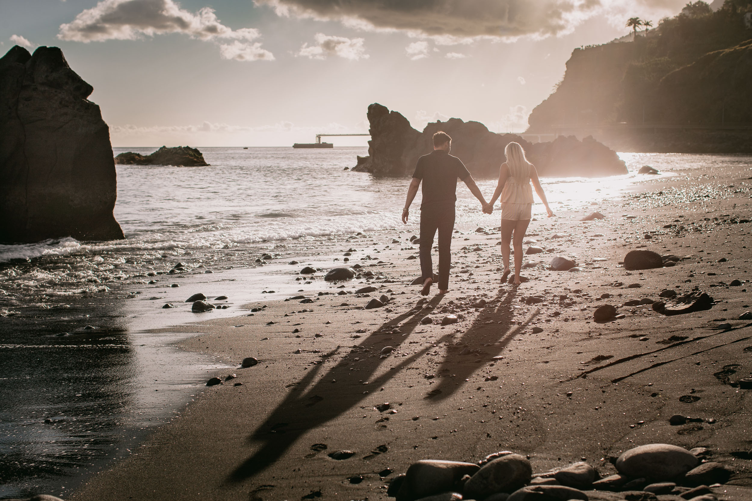 Wide silhouette of Mike and Amelie walking hand in hand into a golden sun on the wet sand, long shadows stretching back toward the camera, sea stack on the left at Praia Formosa, Madeira