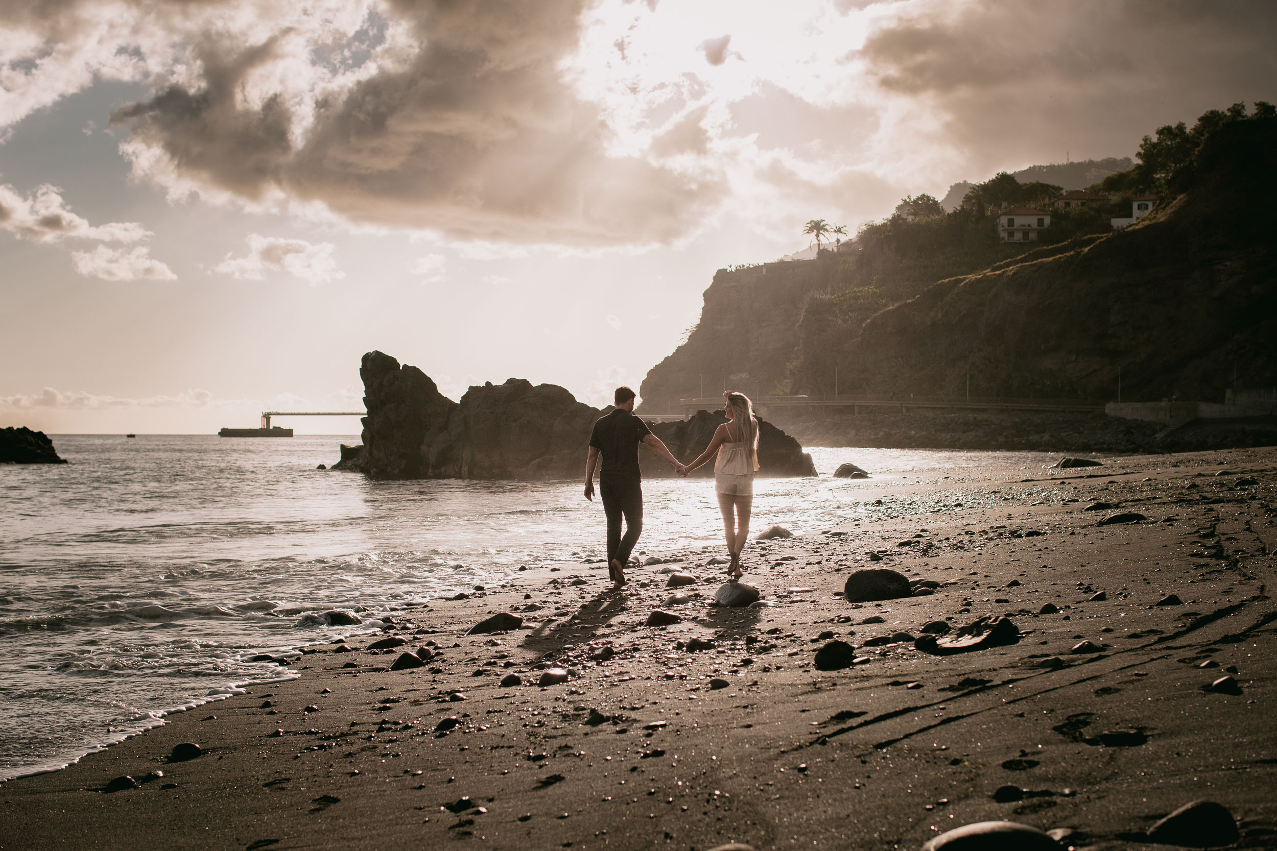 Cinematic wide of Mike and Amelie hand in hand walking toward a low sun, small black sea stacks and the Funchal pier silhouetted against the bright water at Praia Formosa, Madeira