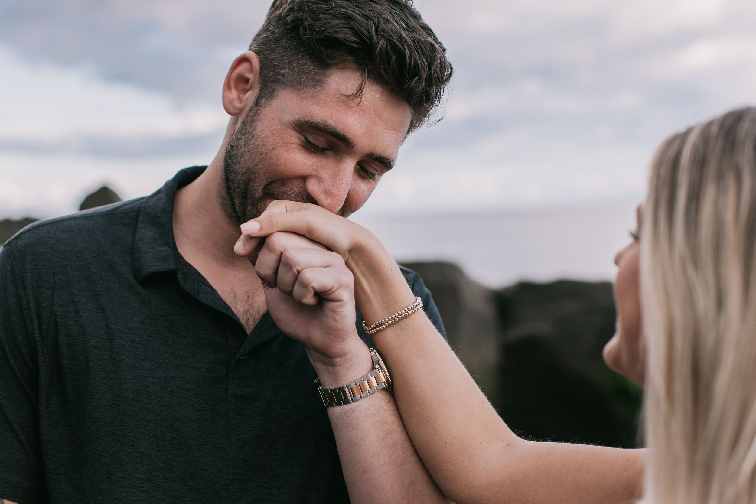 Close portrait of Mike laughing while pressing Amelie's ringed hand to his lips, his silver-and-gold watch on his wrist, soft grey cloudy background at Praia Formosa, Madeira