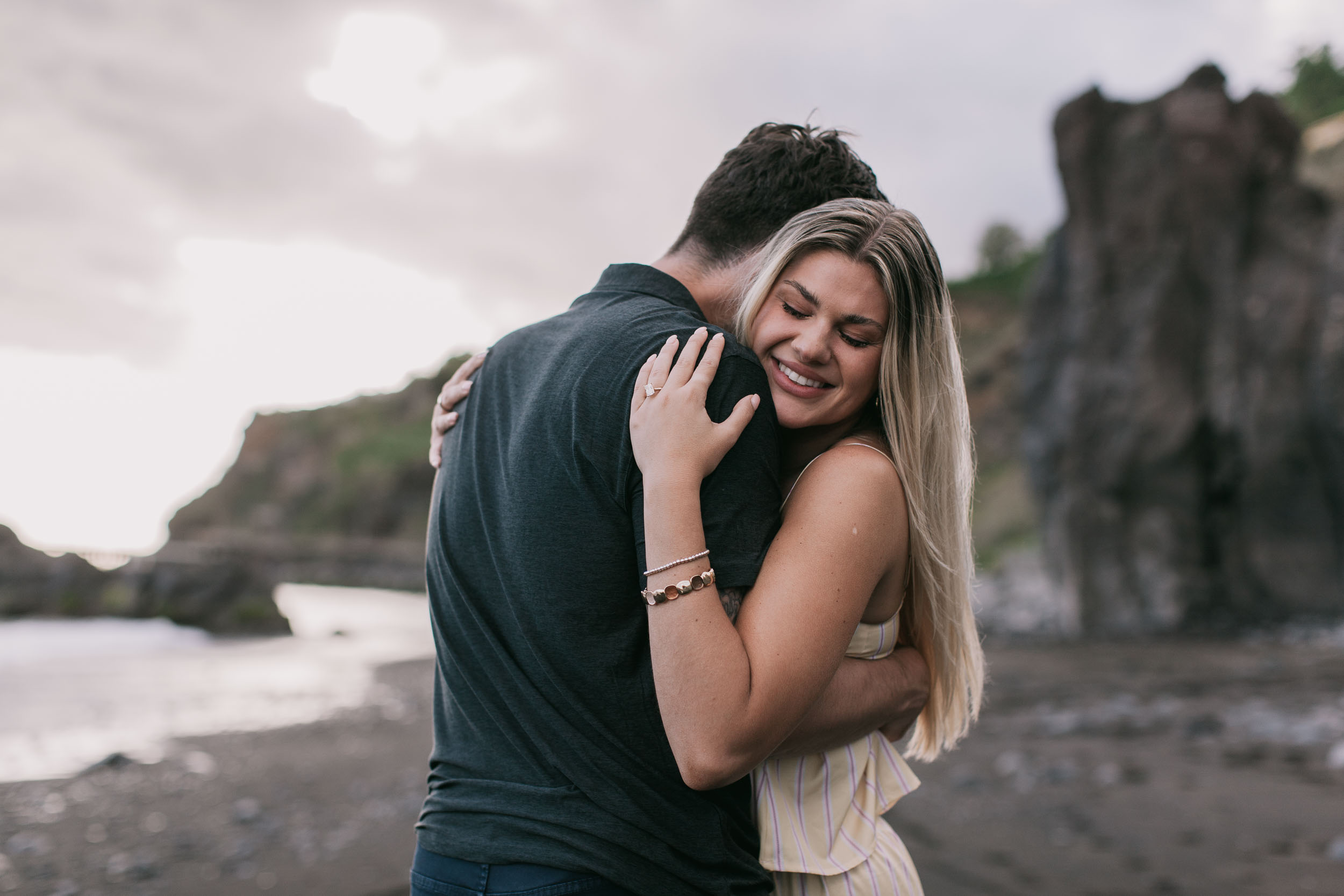 Mike turned away with Amelie's cheek pressed to his shoulder smiling with eyes closed, her ringed hand on his upper arm, dark cliffs and dusk sky behind at Praia Formosa, Madeira
