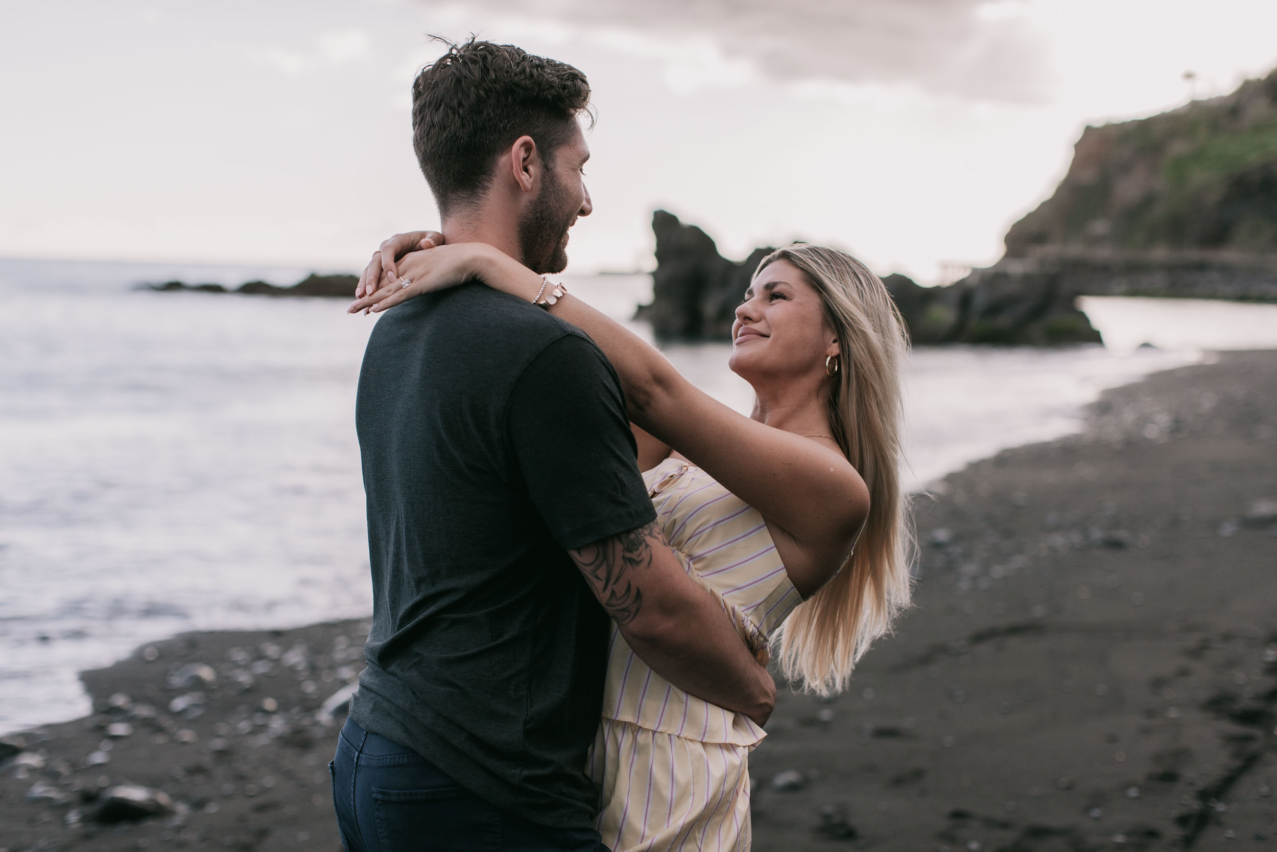 Mike and Amelie standing close holding each other and laughing face to face on the dark sand, his hand on her back and her hand on his neck, sea stack and golden dusk behind at Praia Formosa, Madeira