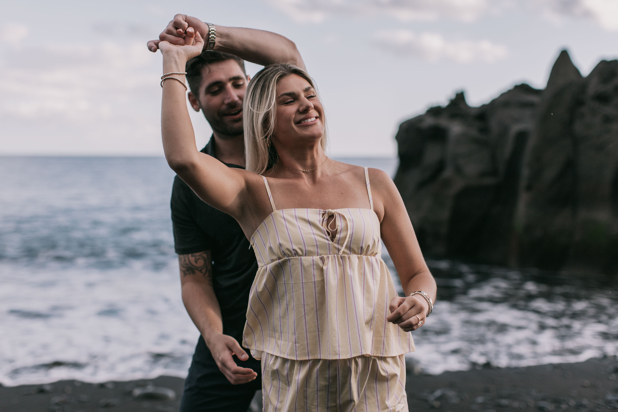 Mike spinning Amelie under his lifted arm at the water's edge, her eyes closed and head tipped back in a wide laugh, dark headland on the right at Praia Formosa, Madeira
