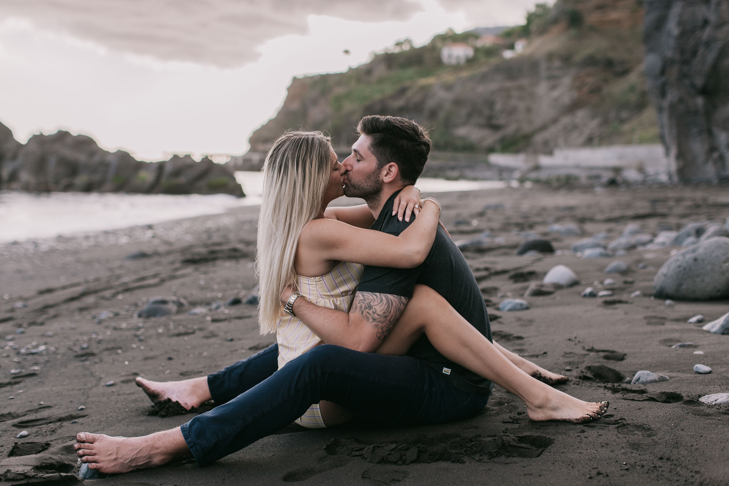 Mike and Amelie sitting cross-legged on the wet black sand kissing, his tattooed arm wrapped around her back, distant Funchal headland visible behind them at Praia Formosa, Madeira