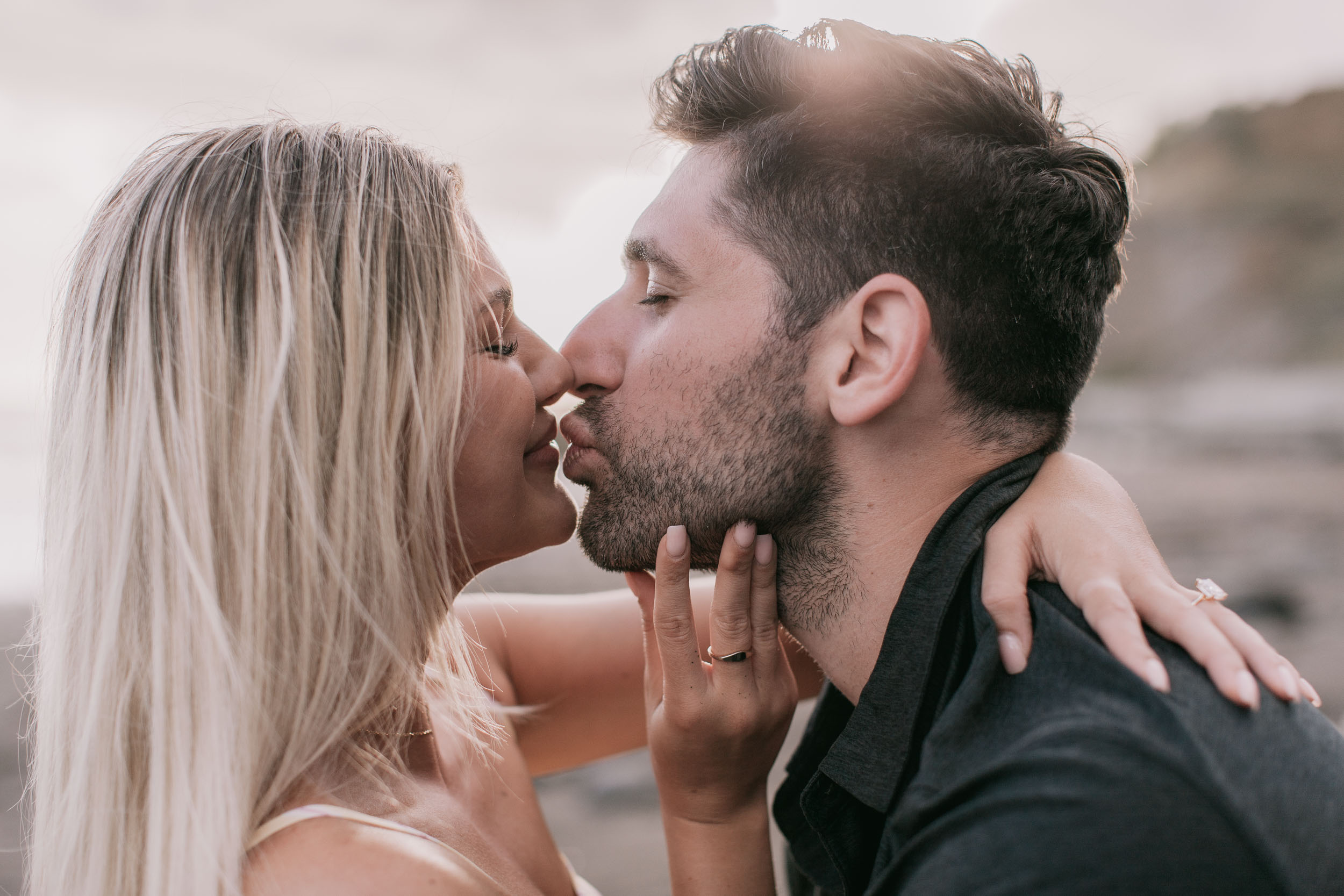 Tight profile of Mike about to kiss Amelie, her ringed fingertips resting on his bearded jaw, gold ring catching the warm late light, Praia Formosa, Madeira