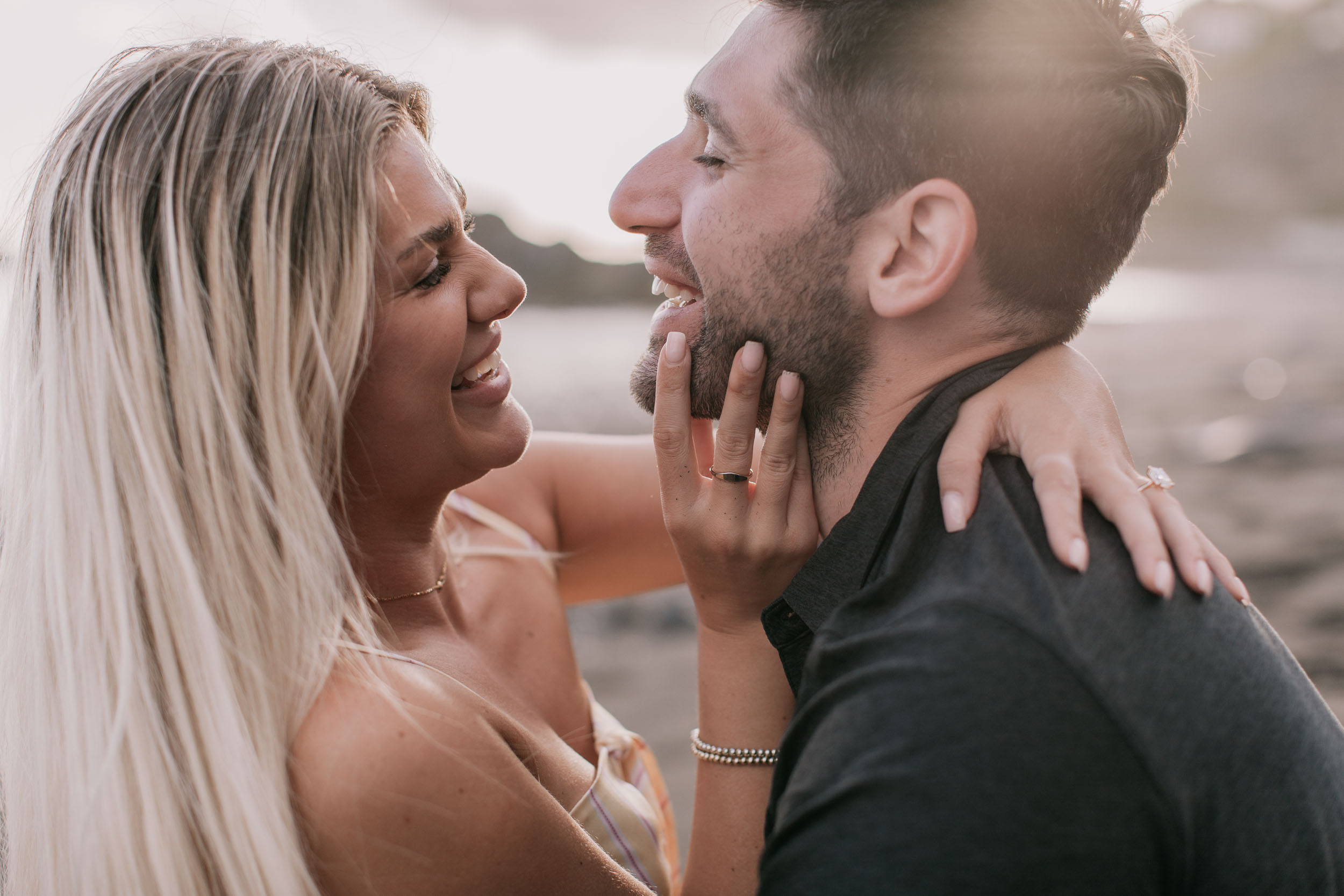 Tight side-profile of Mike and Amelie face to face laughing, her ringed hand cupping his beard, his other ring just visible on his finger at her shoulder, soft warm light at Praia Formosa, Madeira
