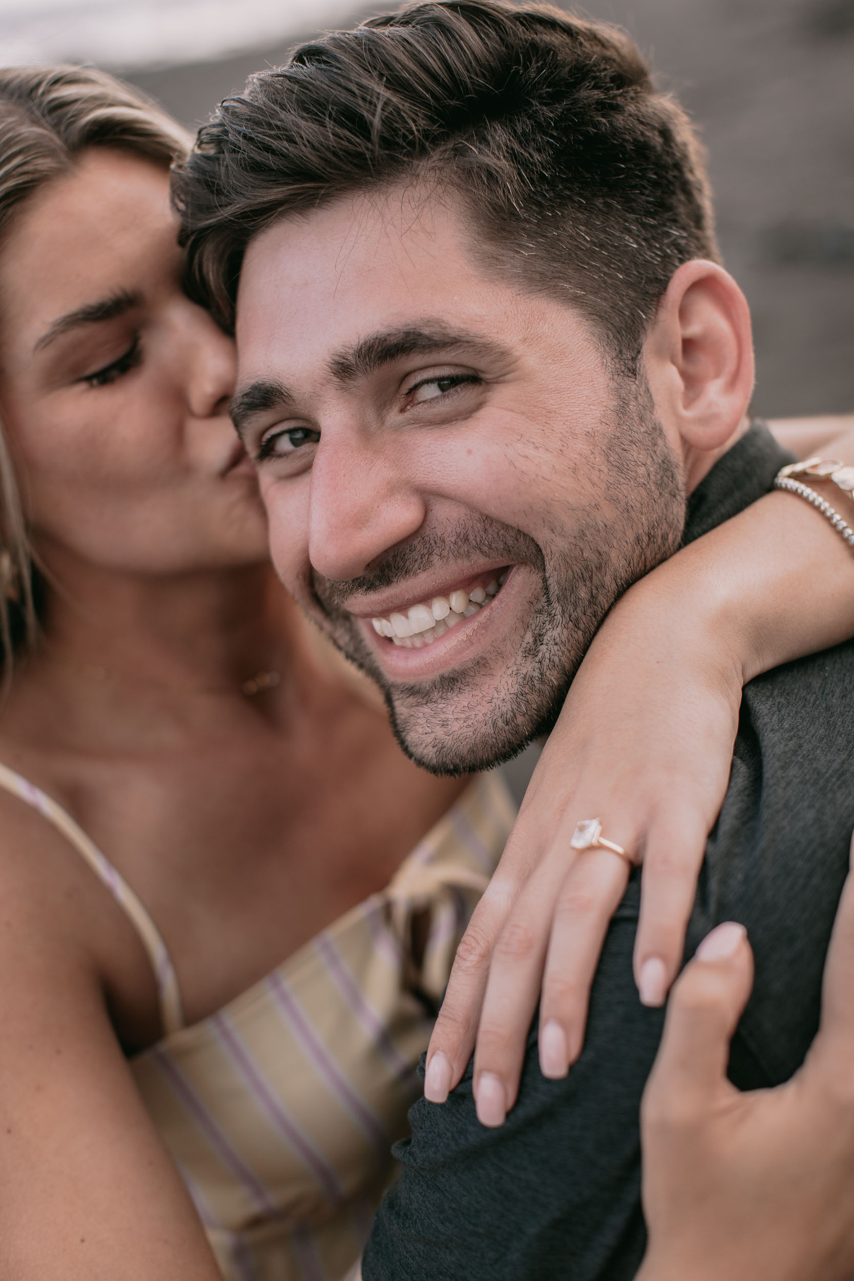 Vertical close-up of Amelie kissing the side of Mike's forehead while he grins straight at the camera, her ringed hand draped across his collar in the foreground, Praia Formosa, Madeira