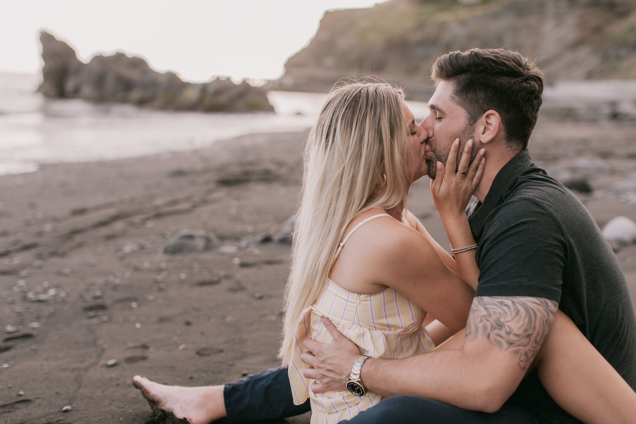 Mike and Amelie sitting facing each other kissing on the wet sand, his tattooed forearm and watch resting on her thigh, small sea stack on the left at Praia Formosa, Madeira
