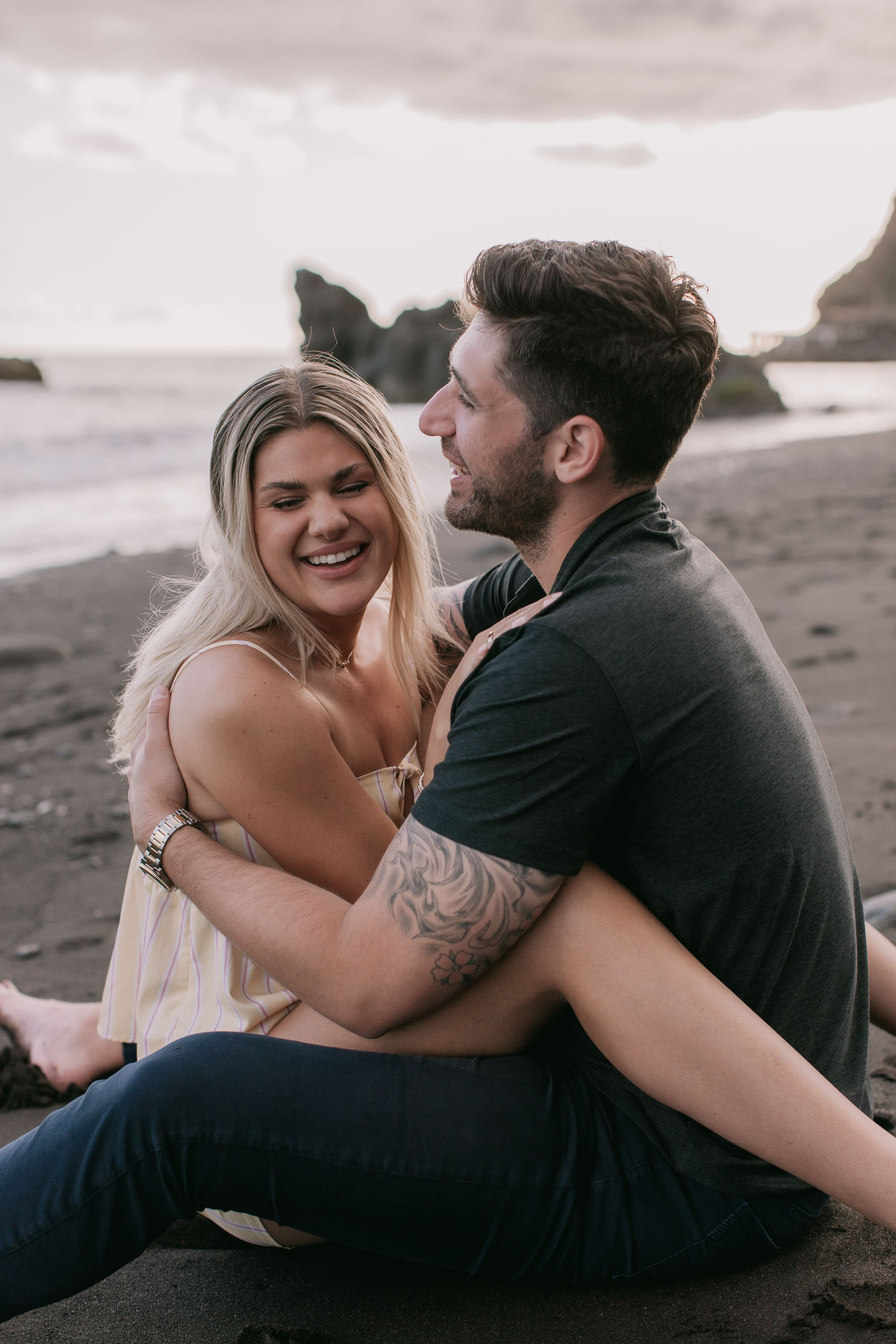 Mike and Amelie sitting on the dark sand laughing nose to nose, his arms wrapped under her legs and the tribal sleeve tattoo curling around her thigh, Praia Formosa, Madeira