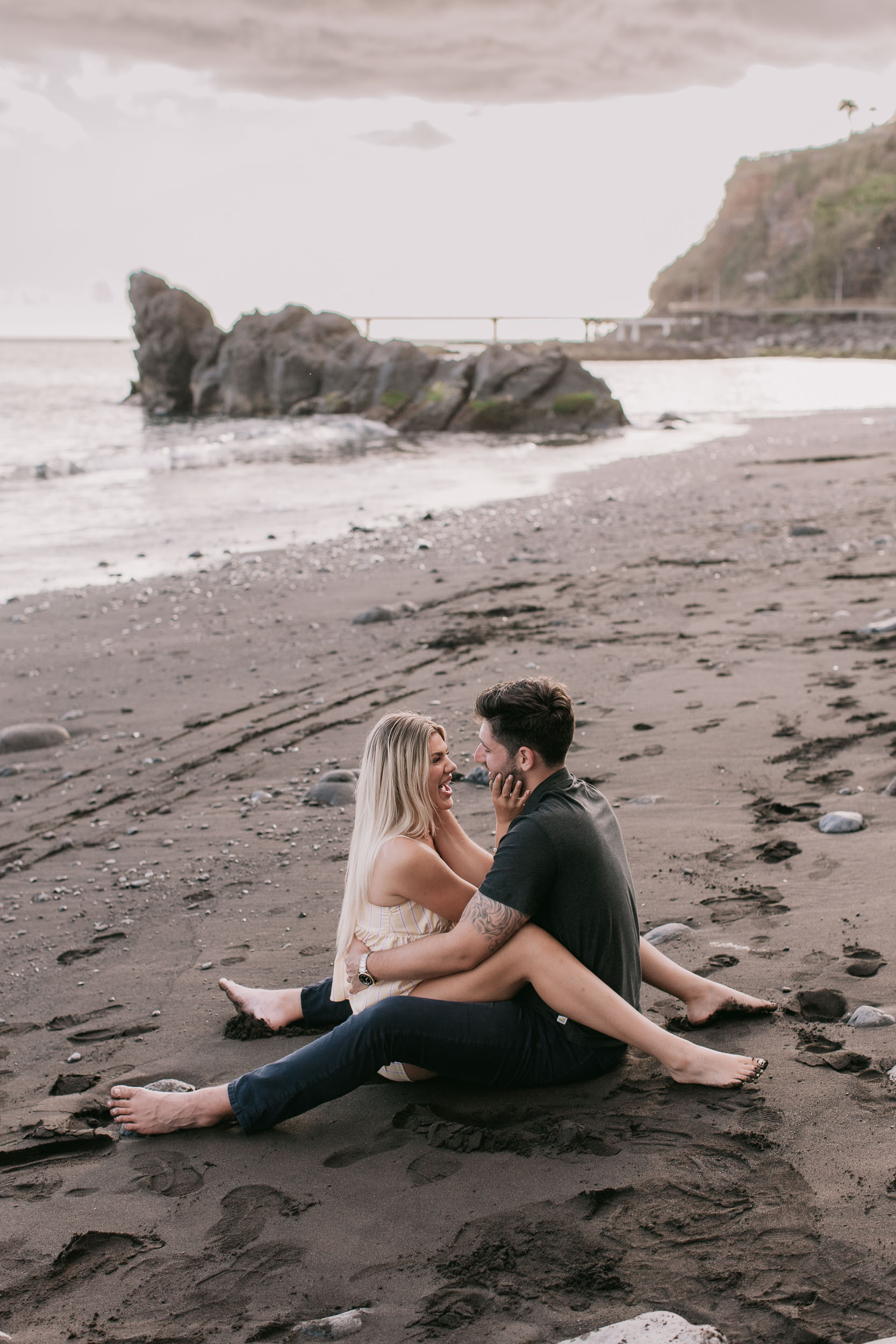 Wide frame of Mike and Amelie sitting facing each other on the wet sand with hands on each other's faces laughing, a curved sea stack rising from the water behind them at Praia Formosa, Madeira