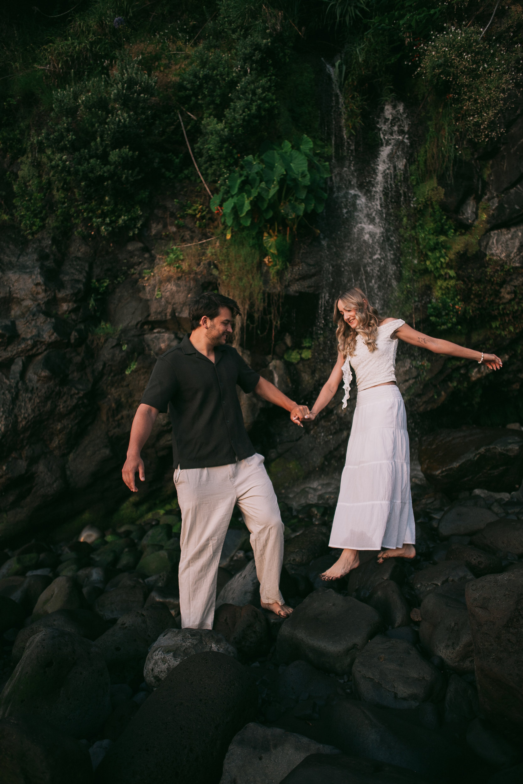 She spreads an arm wide on the waterfall rocks, holding his hand, Seixal, Madeira