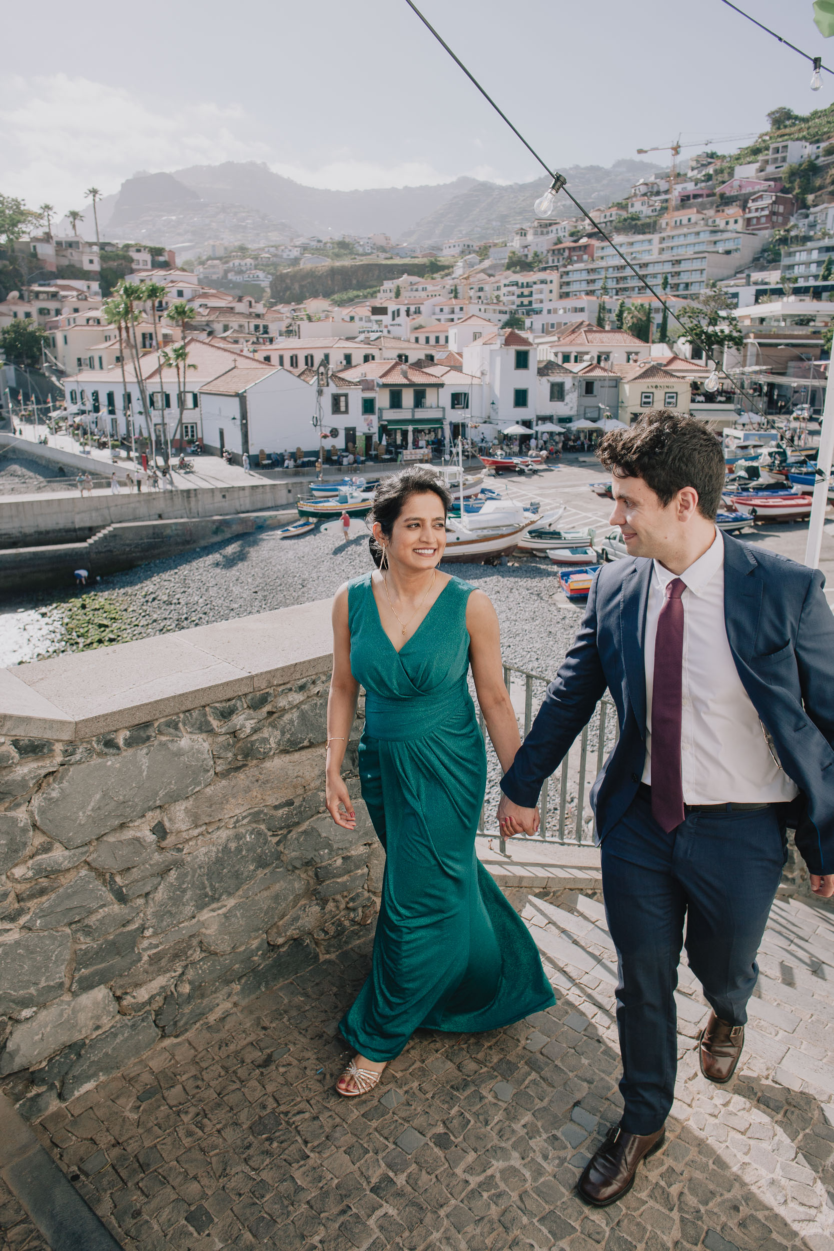 Couple climbing a stone staircase hand in hand, the harbour and fishing boats of Câmara de Lobos below, Madeira