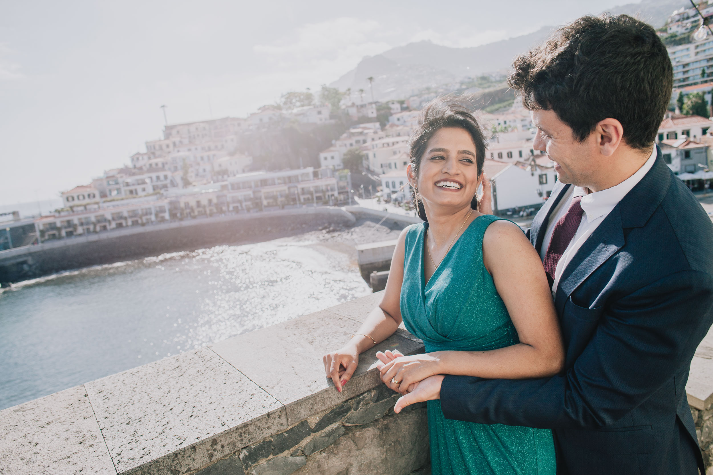 Bride laughing as she leans on a stone wall beside the groom, Câmara de Lobos village rising behind, Madeira