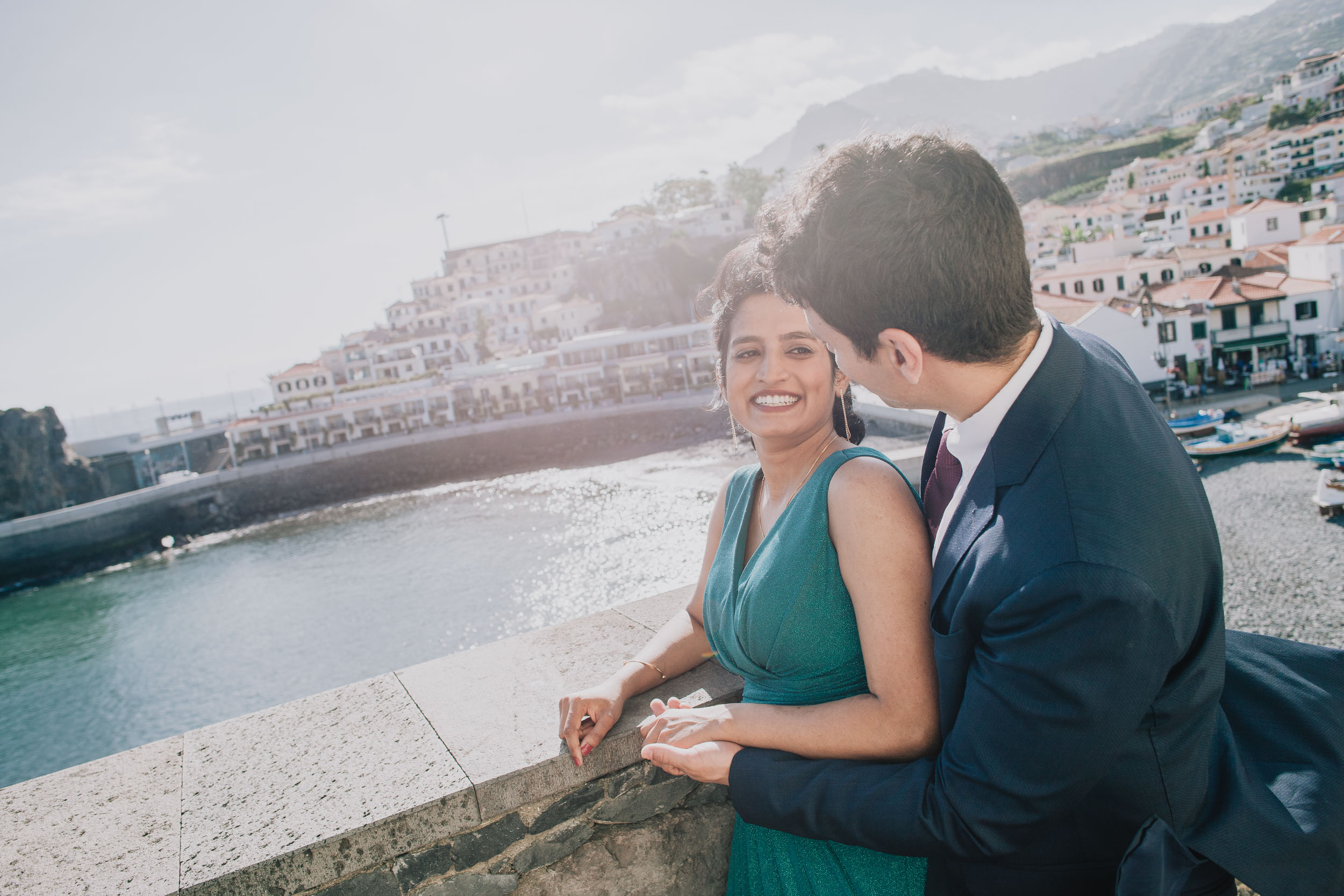 She laughs looking out over the harbour while he leans in behind her, their hands together on the stone wall, Camara de Lobos, Madeira