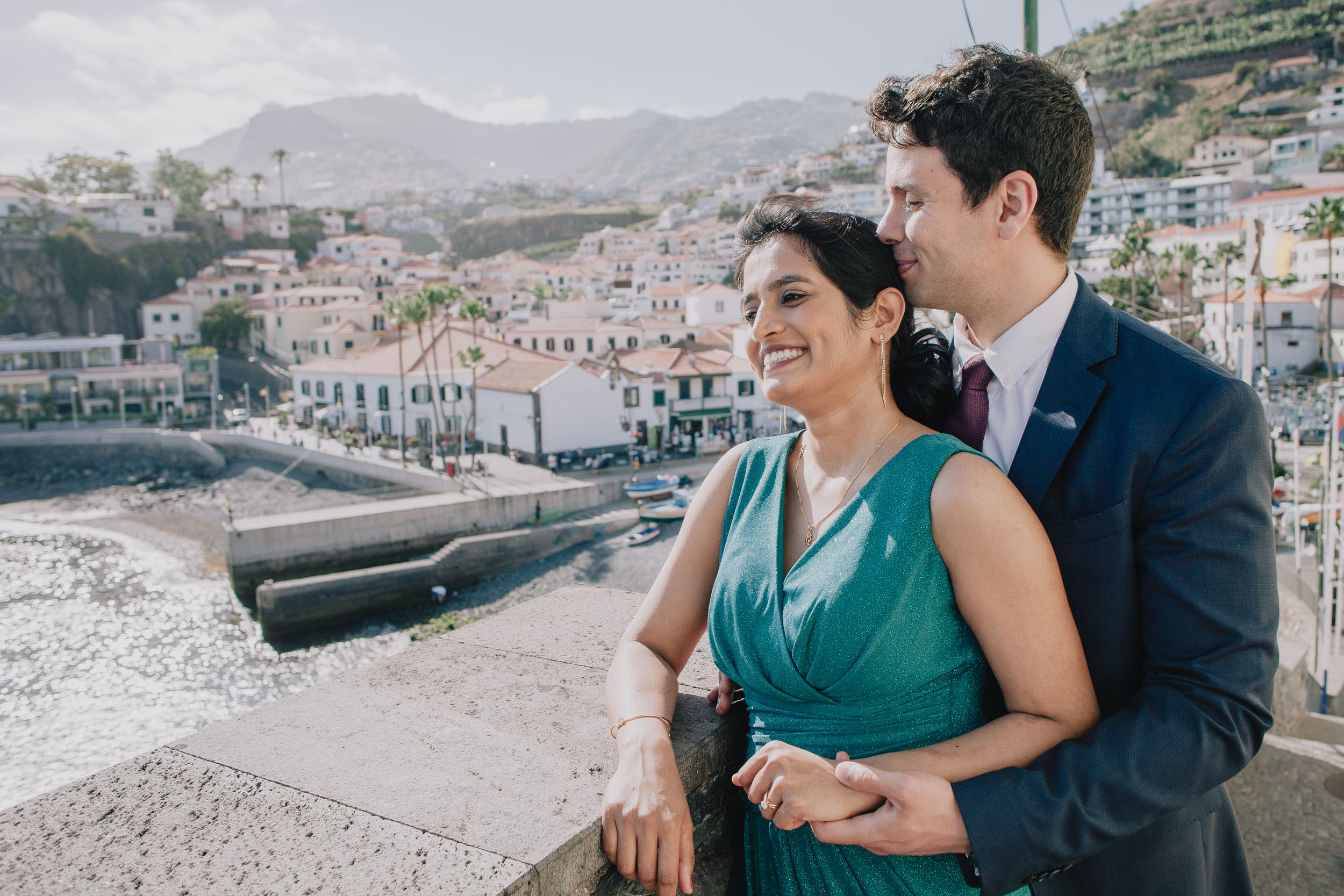 Groom embracing the bride from behind as they look out over the full sweep of Câmara de Lobos harbour and village, Madeira
