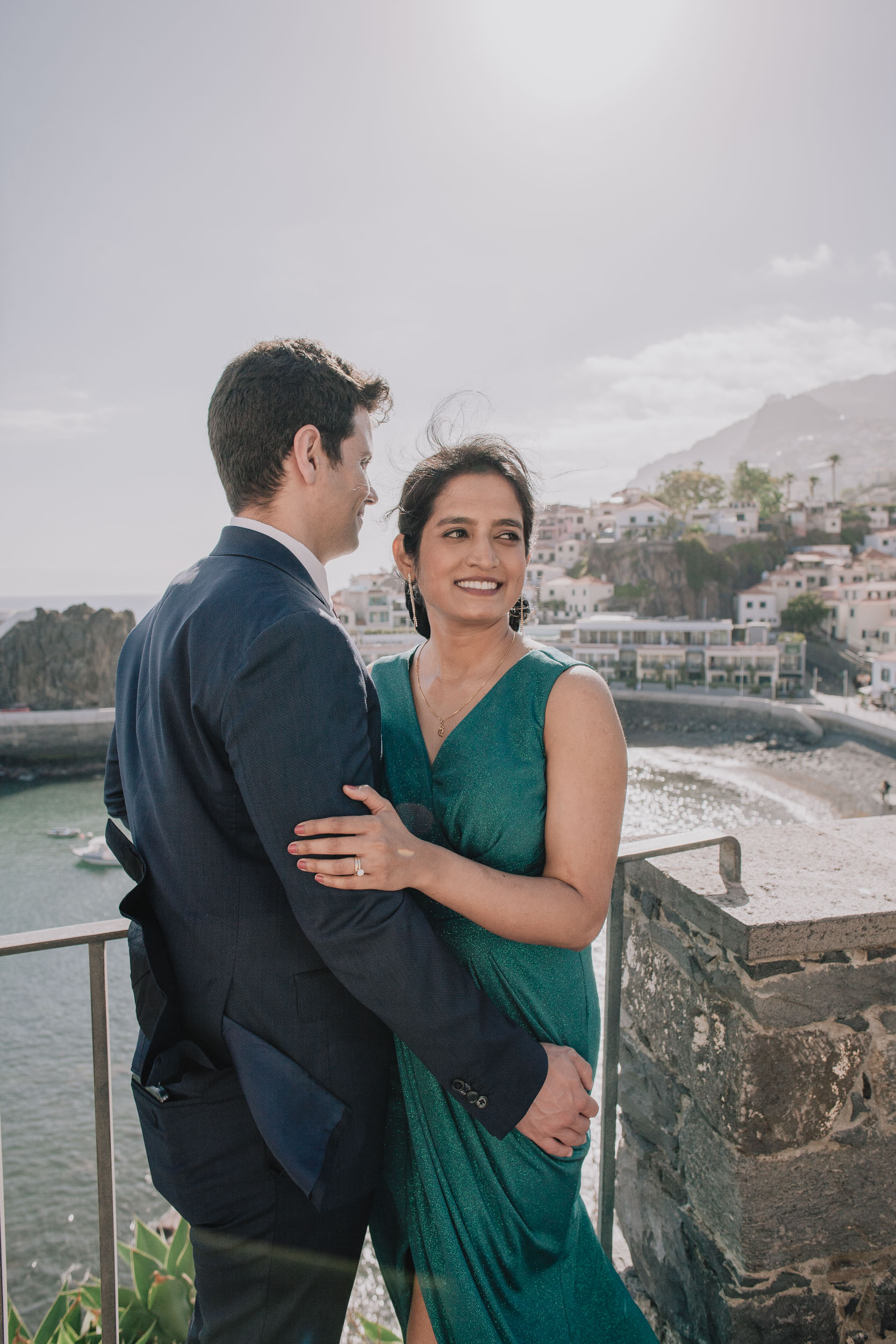 Couple embracing at a railing, the bride smiling at the camera, Câmara de Lobos village behind, Madeira