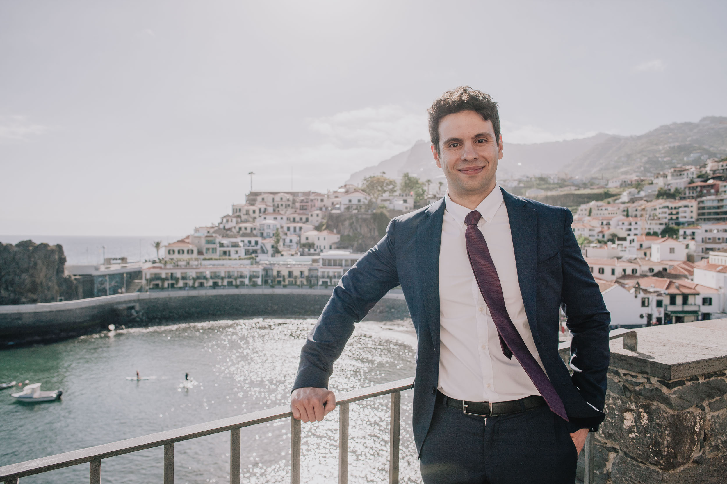 Solo portrait of him at the railing in a navy suit, the harbour and the village on the slope behind, Camara de Lobos, Madeira