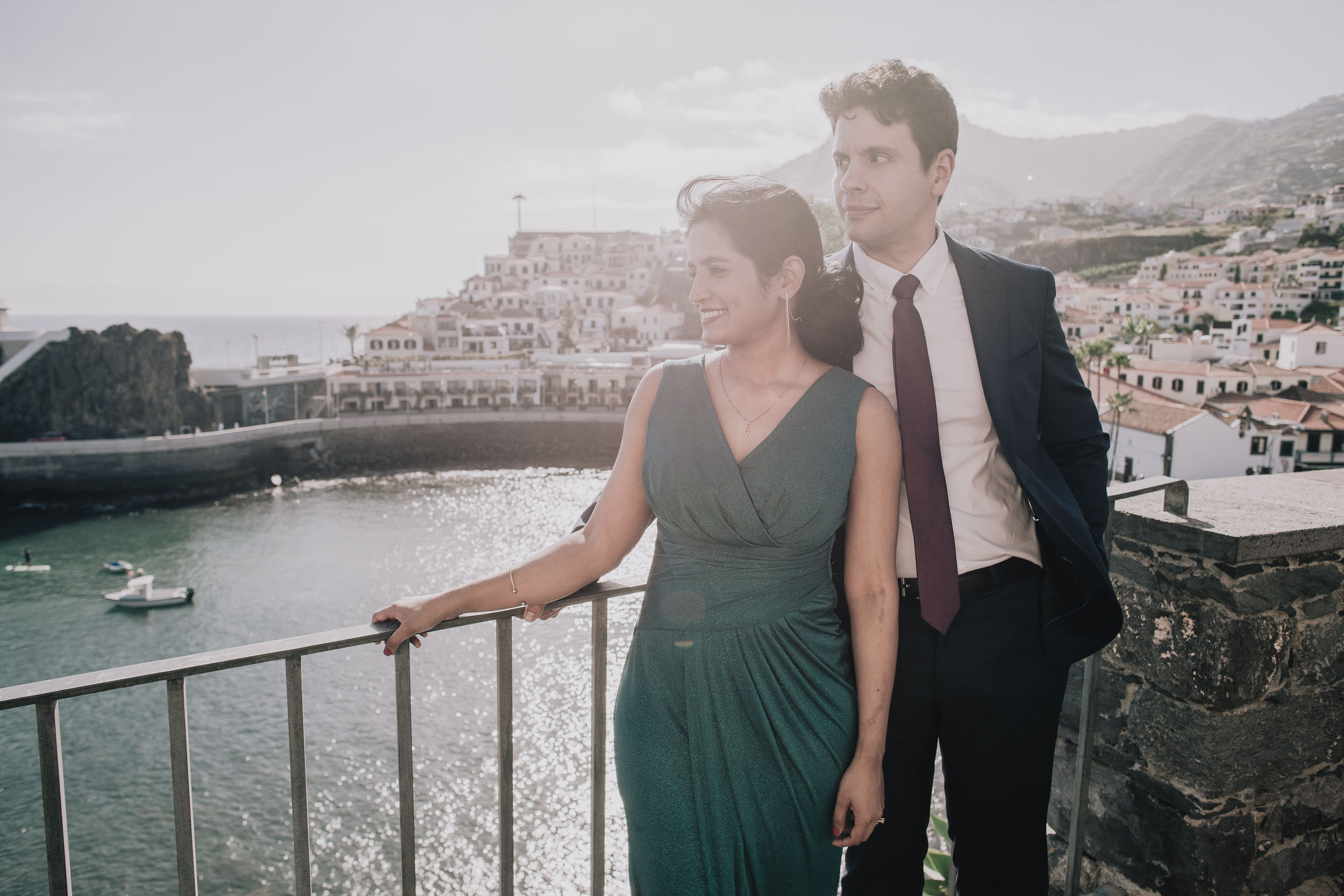 Couple standing at a railing looking out over Câmara de Lobos harbour and the mountains beyond, Madeira