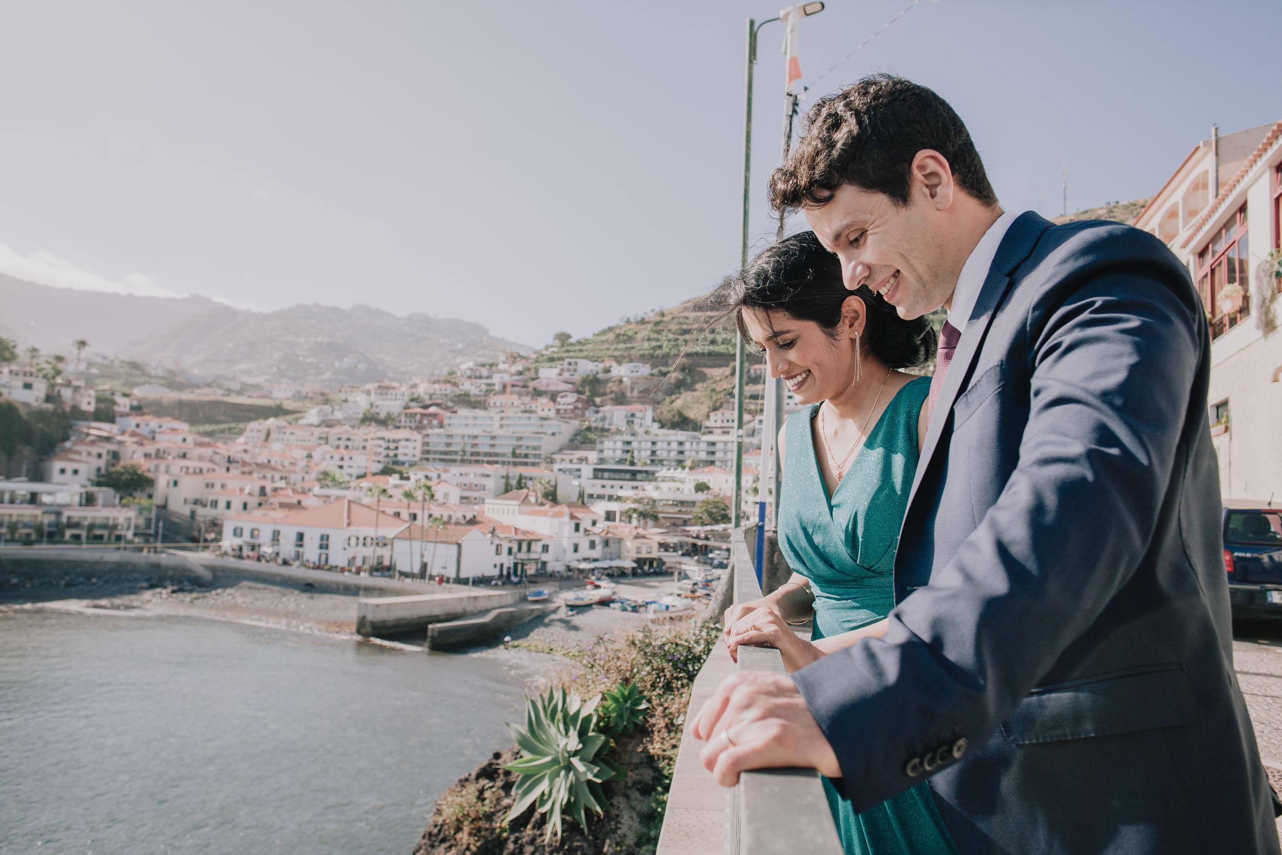 Both lean on the wall looking down at the water, smiling, the village and the terraced hills across the bay, Camara de Lobos, Madeira
