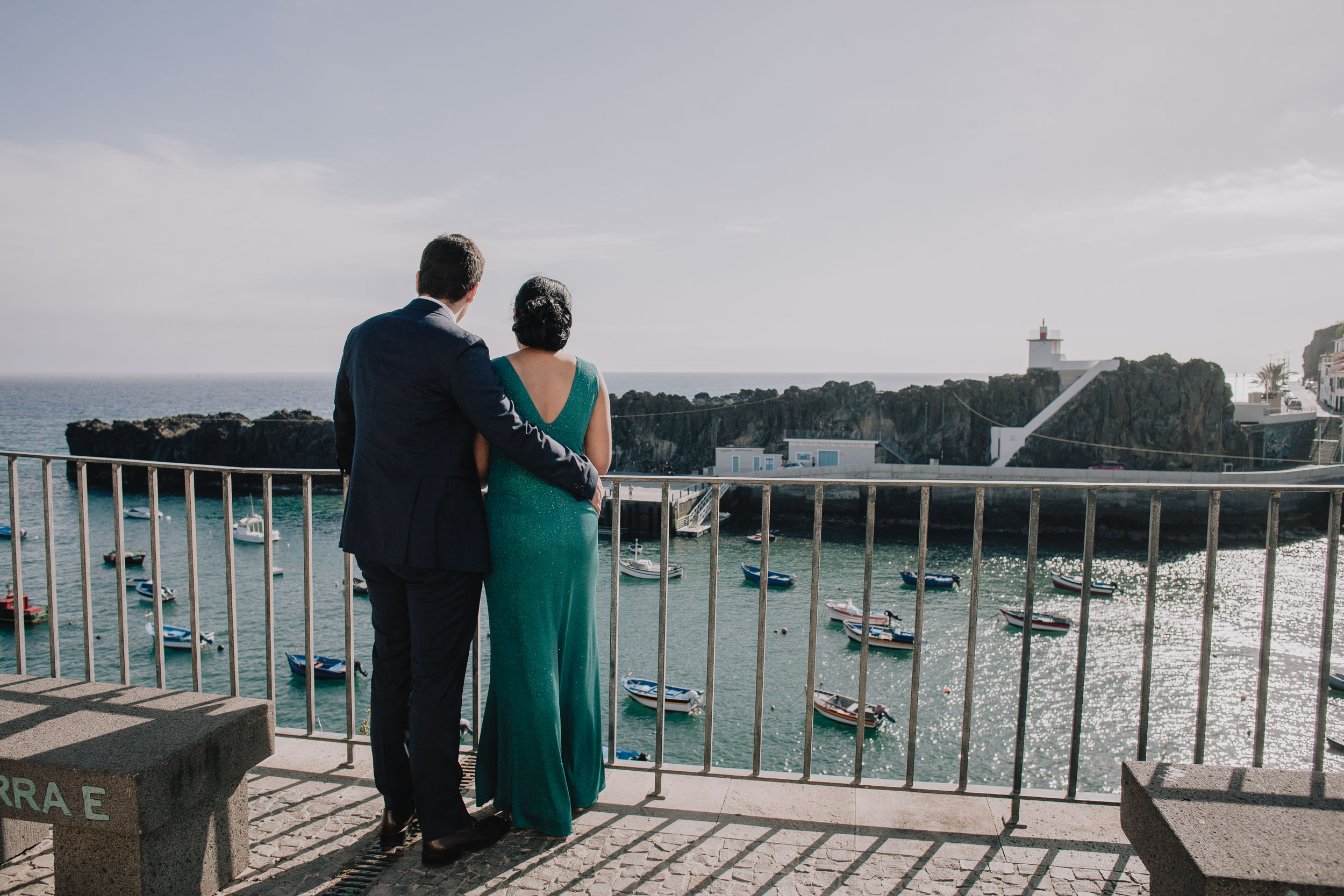 Seen from behind, they stand at the railing with his arm around her and look out at the boats and the lighthouse, Camara de Lobos, Madeira