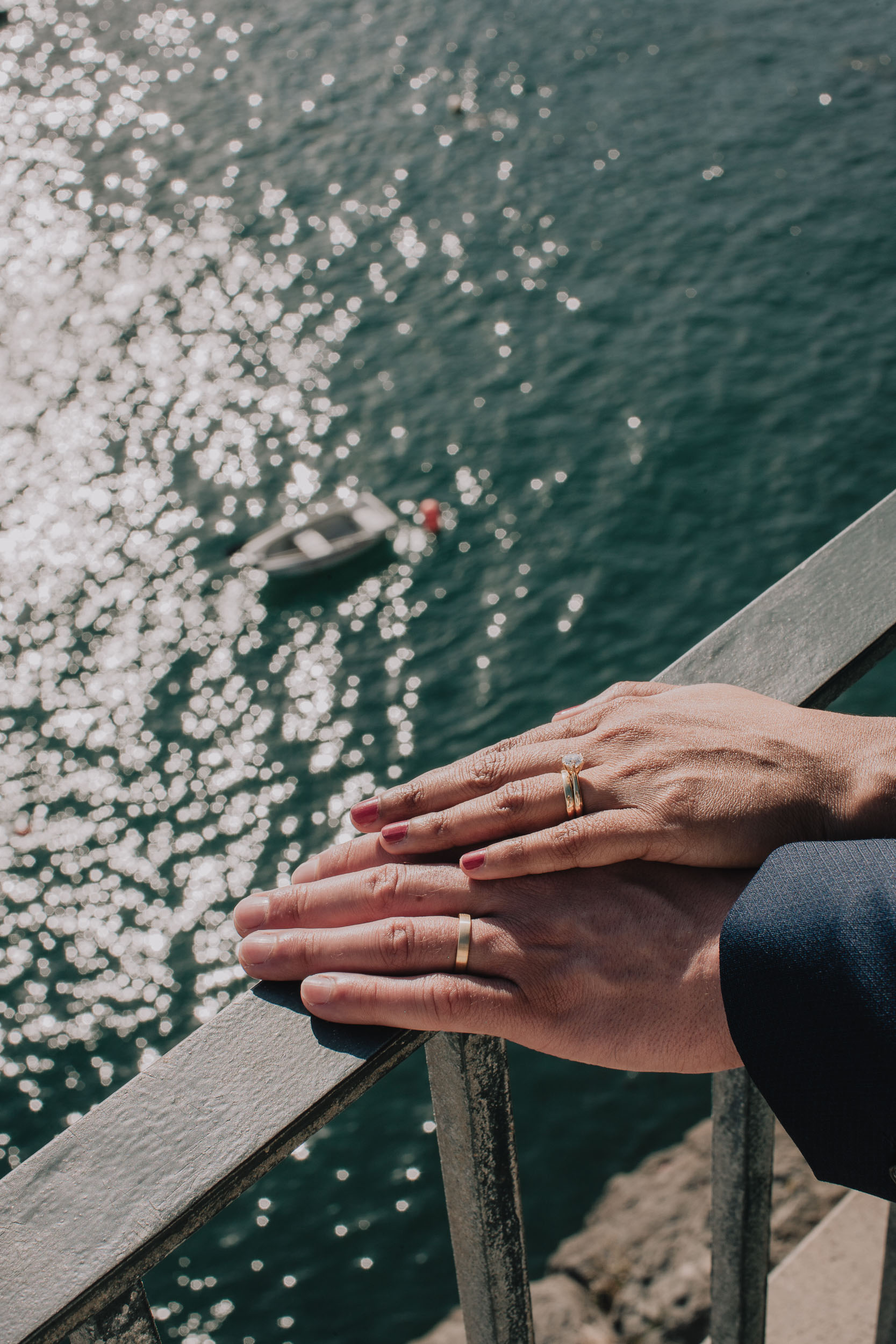 Detail. Their hands one on the other on the railing, a single fishing boat far below on the green water, Camara de Lobos, Madeira