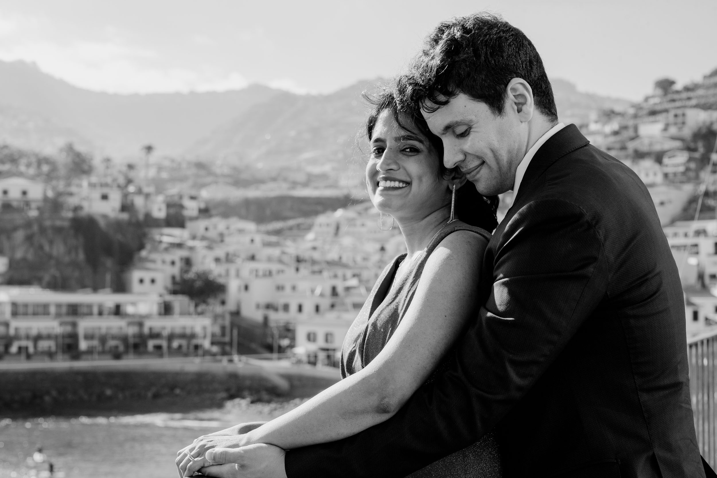 Black and white portrait of the couple embracing, the bride smiling, Câmara de Lobos village behind, Madeira